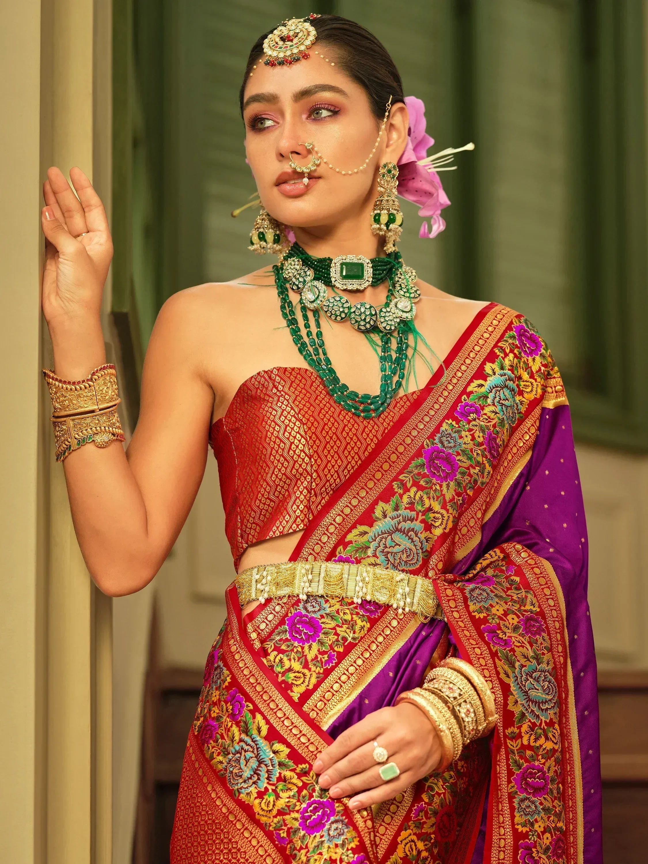 Woman in red and purple floral saree with gold embroidery, green jewelry, and traditional Indian bridal makeup