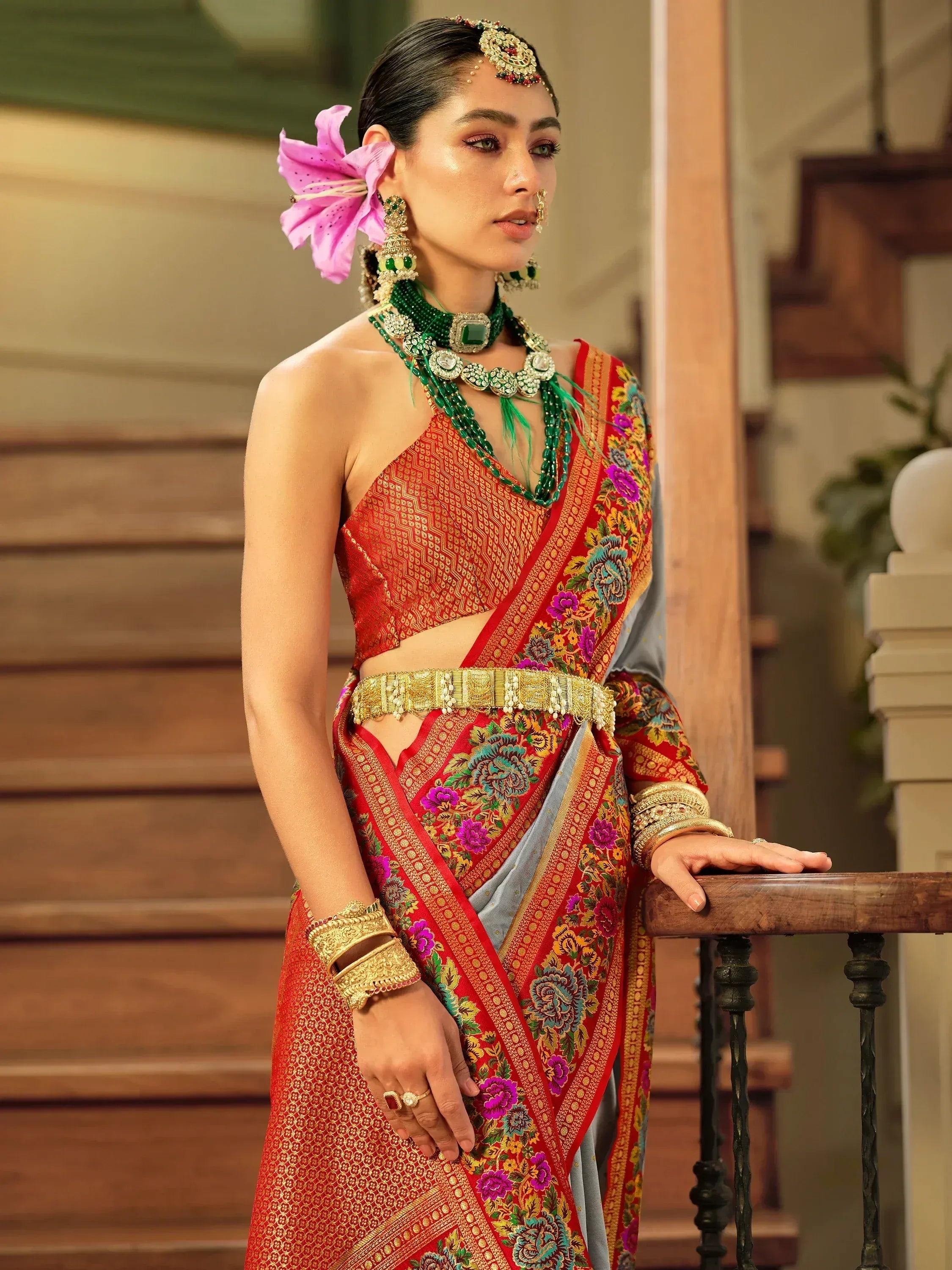 Woman in red floral saree with gold embroidery, green jewelry, and pink flower hair accessory