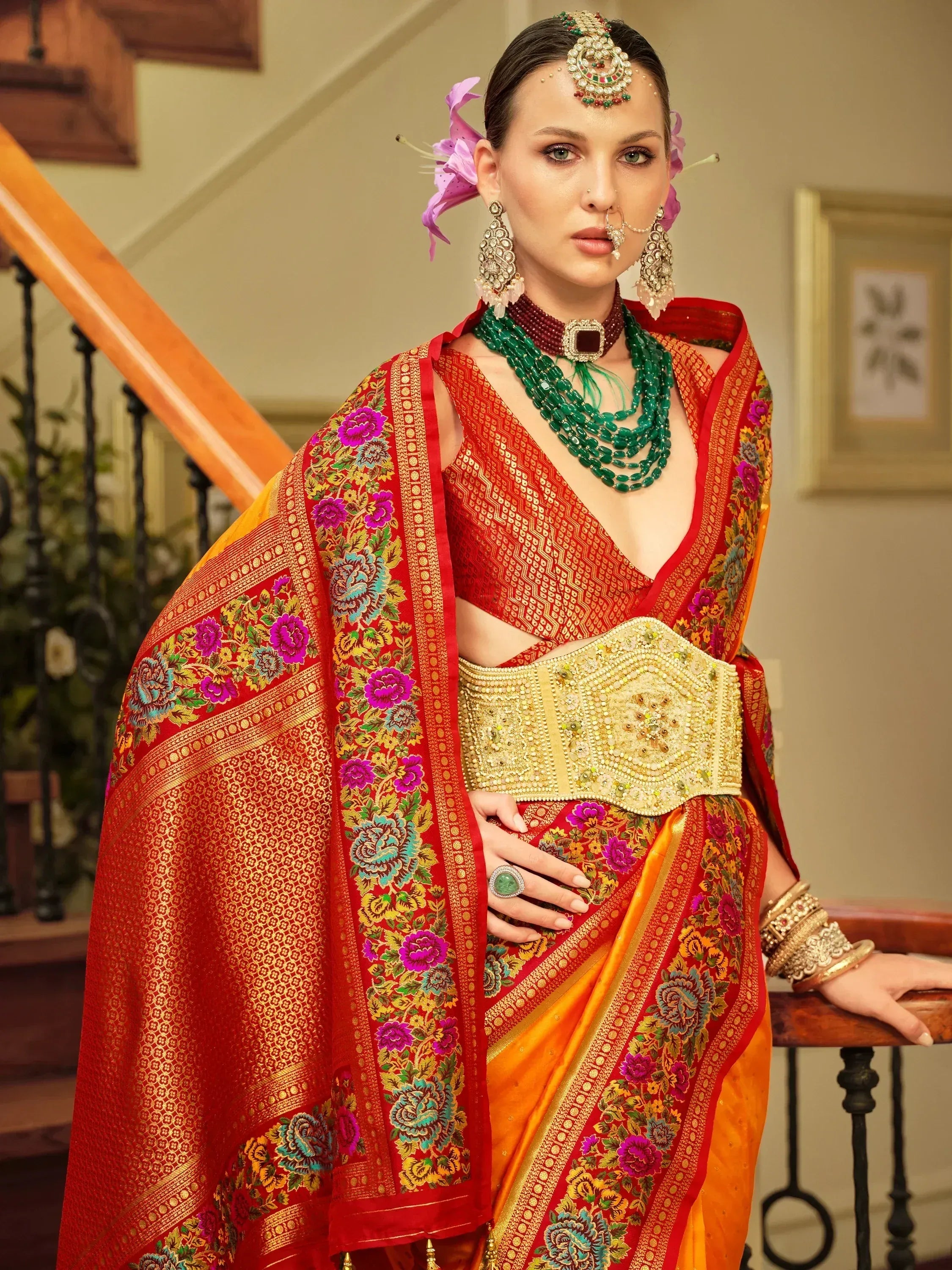 Red and orange floral saree with gold embroidery, paired with traditional Indian jewelry and a decorative belt.