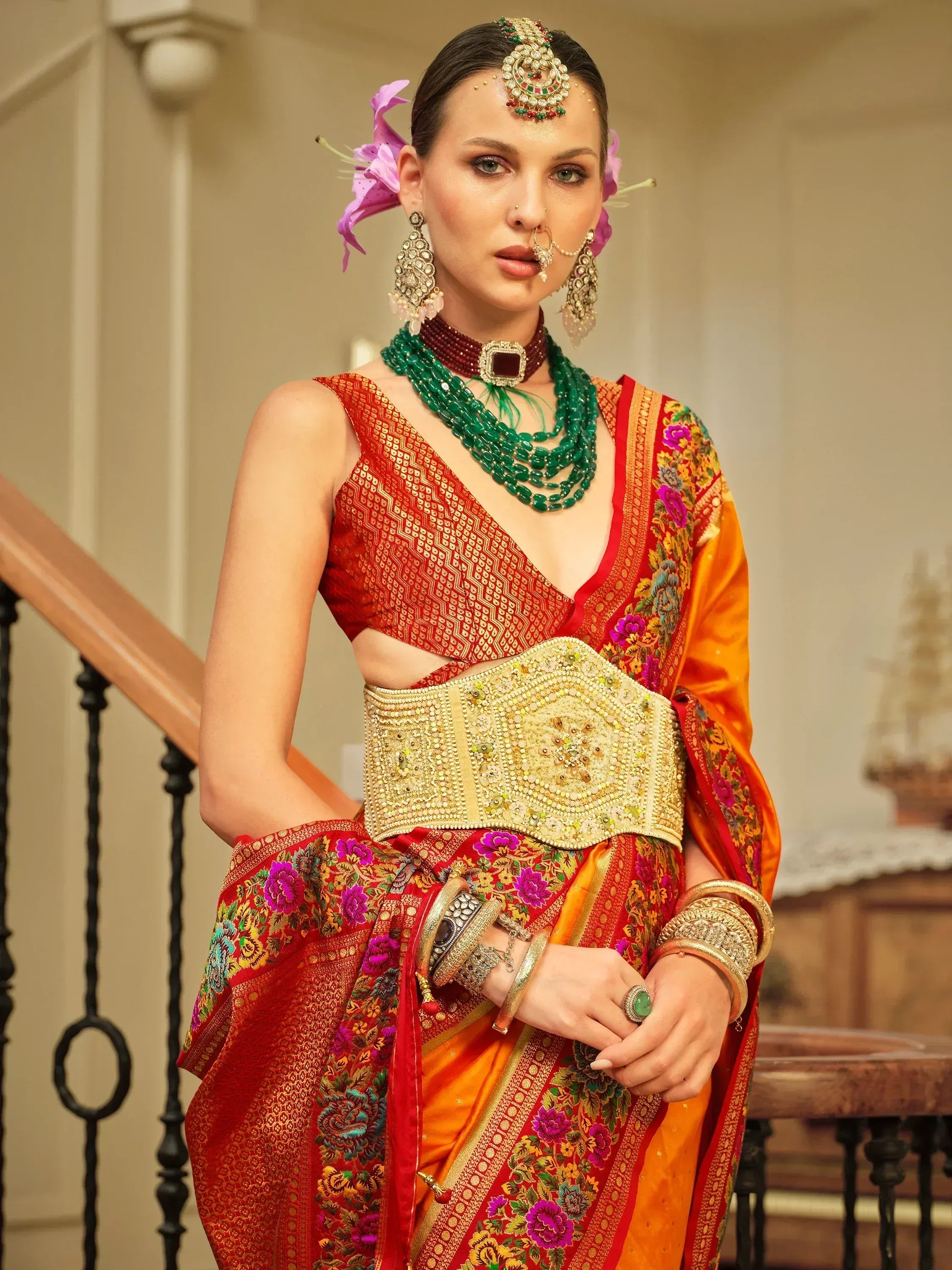 Woman in vibrant red and orange embroidered saree with gold belt, traditional Indian jewelry, and floral hair accessories.