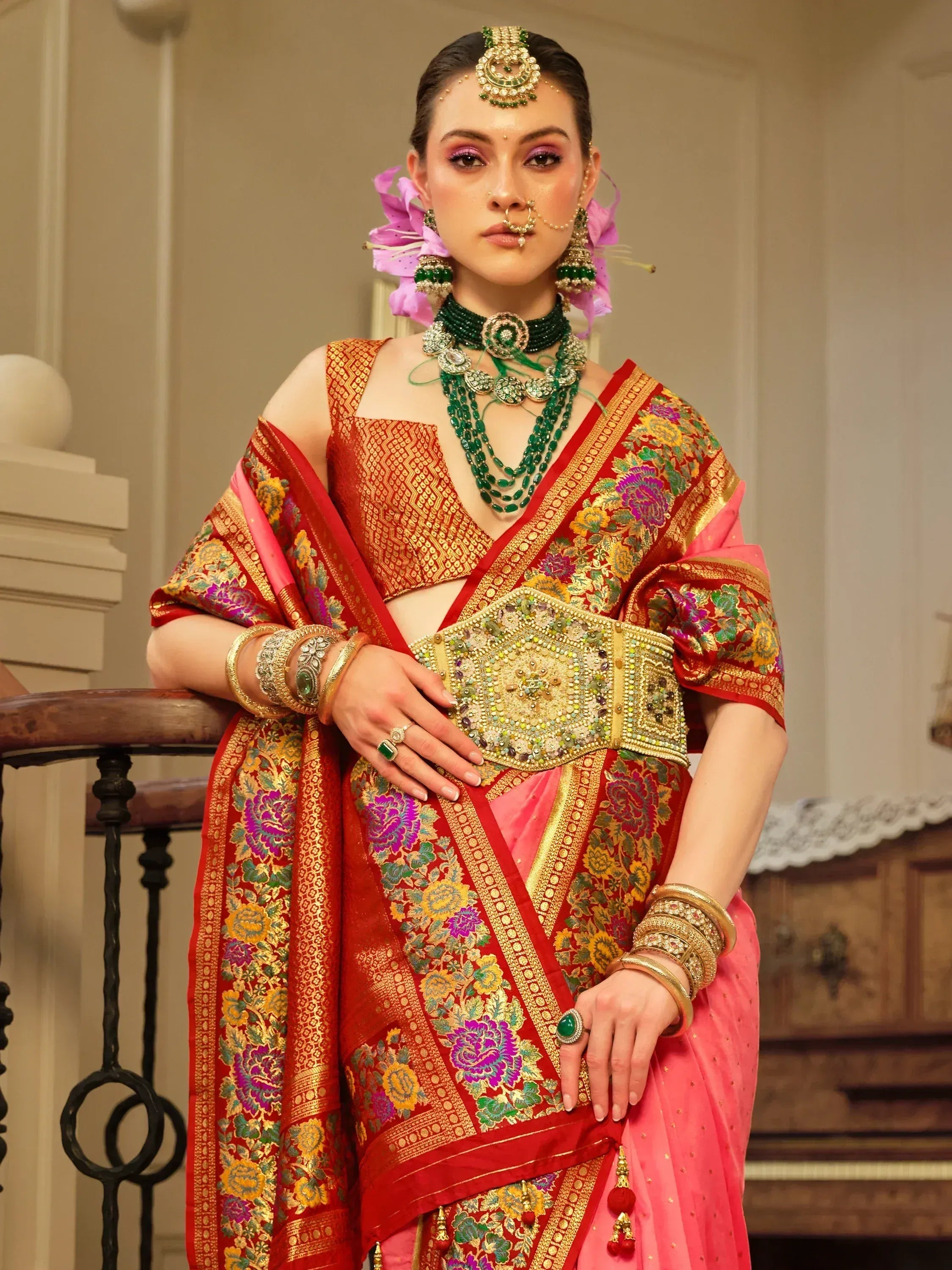 Woman in red and pink floral saree with gold embroidery, green jewelry, and traditional Indian bridal accessories