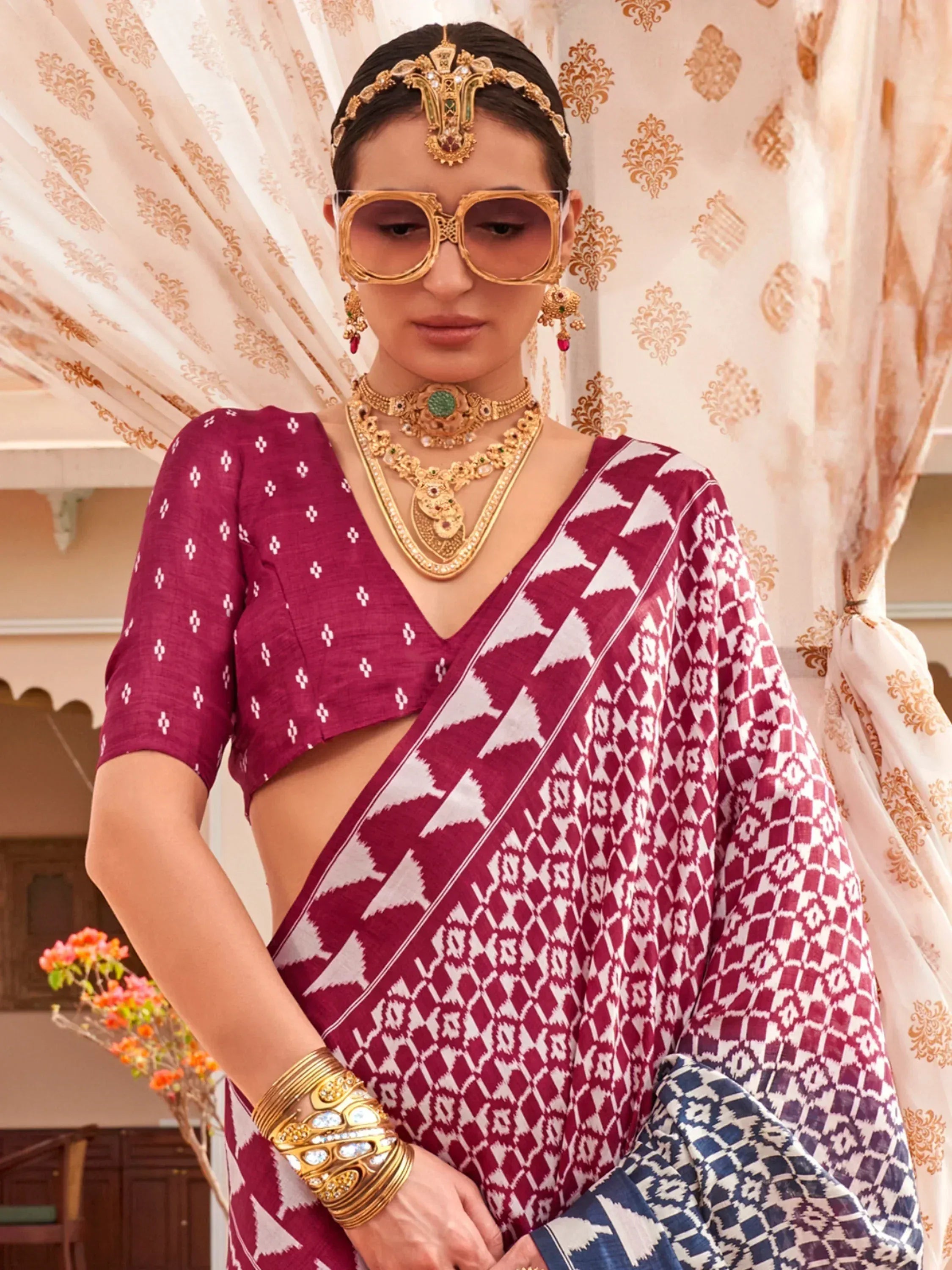 Woman in maroon and white ikat saree with gold jewelry and statement sunglasses, ethnic Indian wear
