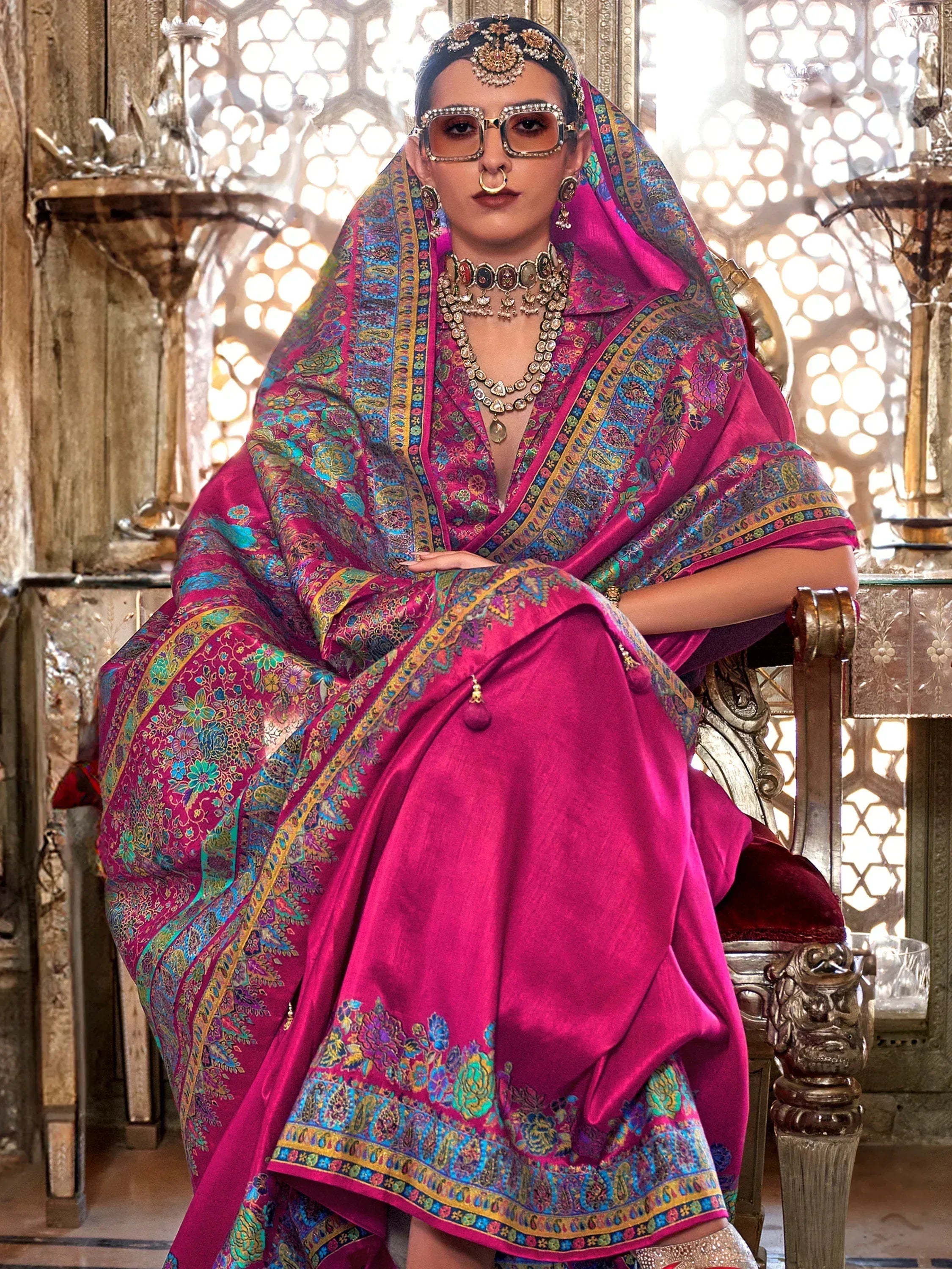 Woman in vibrant pink ethnic saree with intricate floral paisley border, traditional jewelry, and ornate setting
