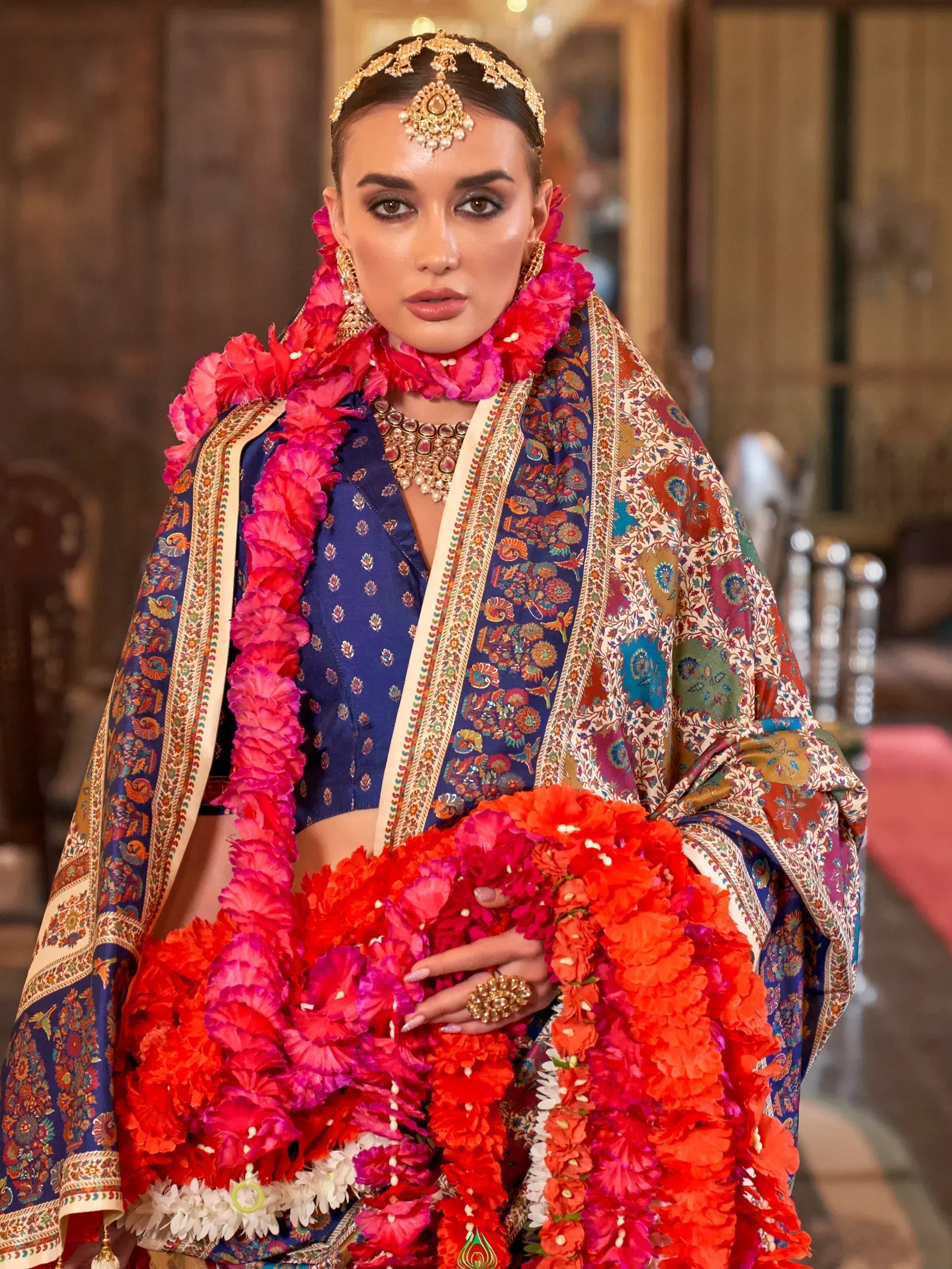 Woman in royal blue embroidered lehenga choli with floral dupatta, gold jewelry, and vibrant flower garlands for Indian wedding.