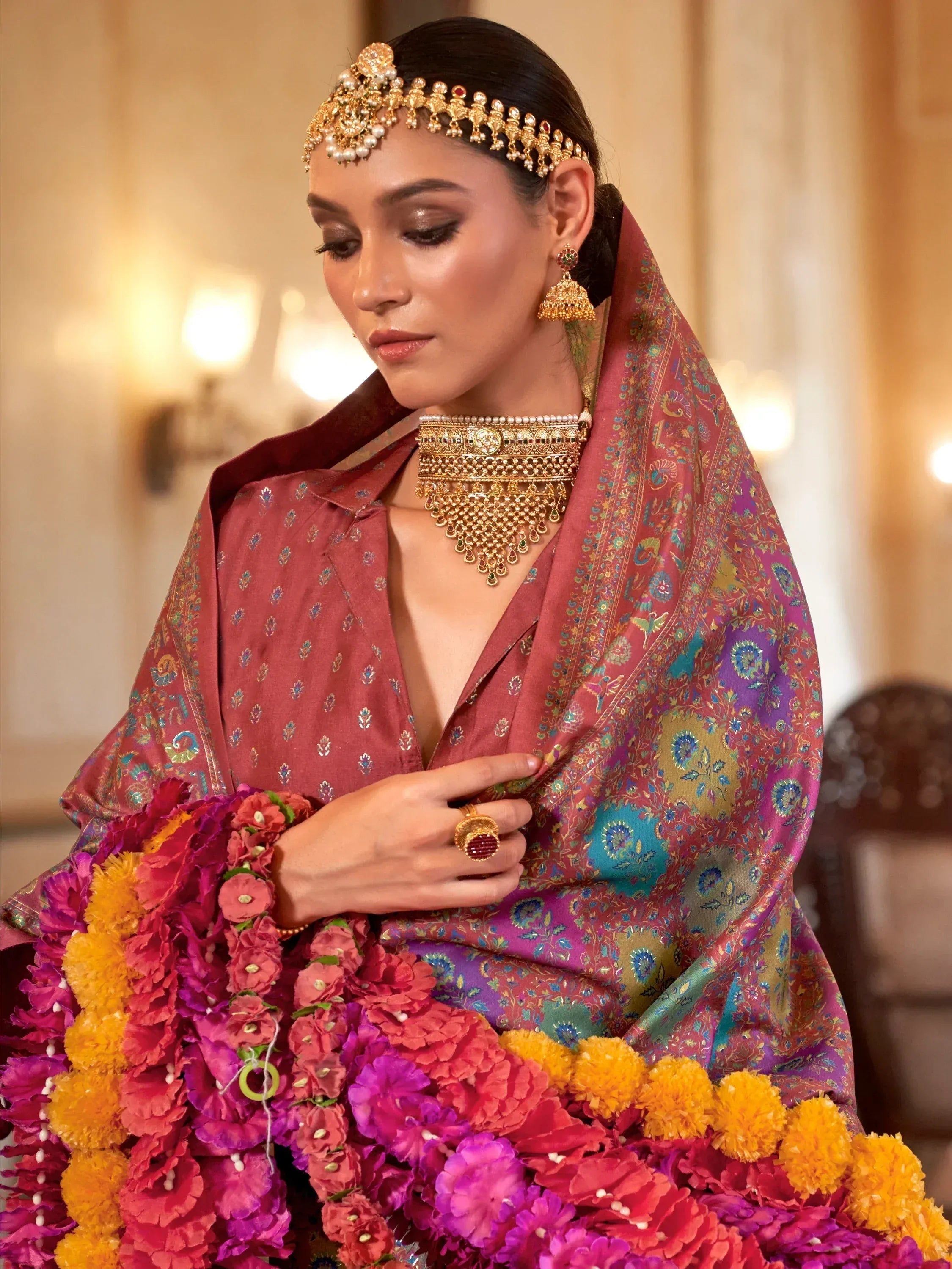 Elegant woman in maroon ethnic saree with gold jewelry, floral garland, and traditional Indian headpiece.