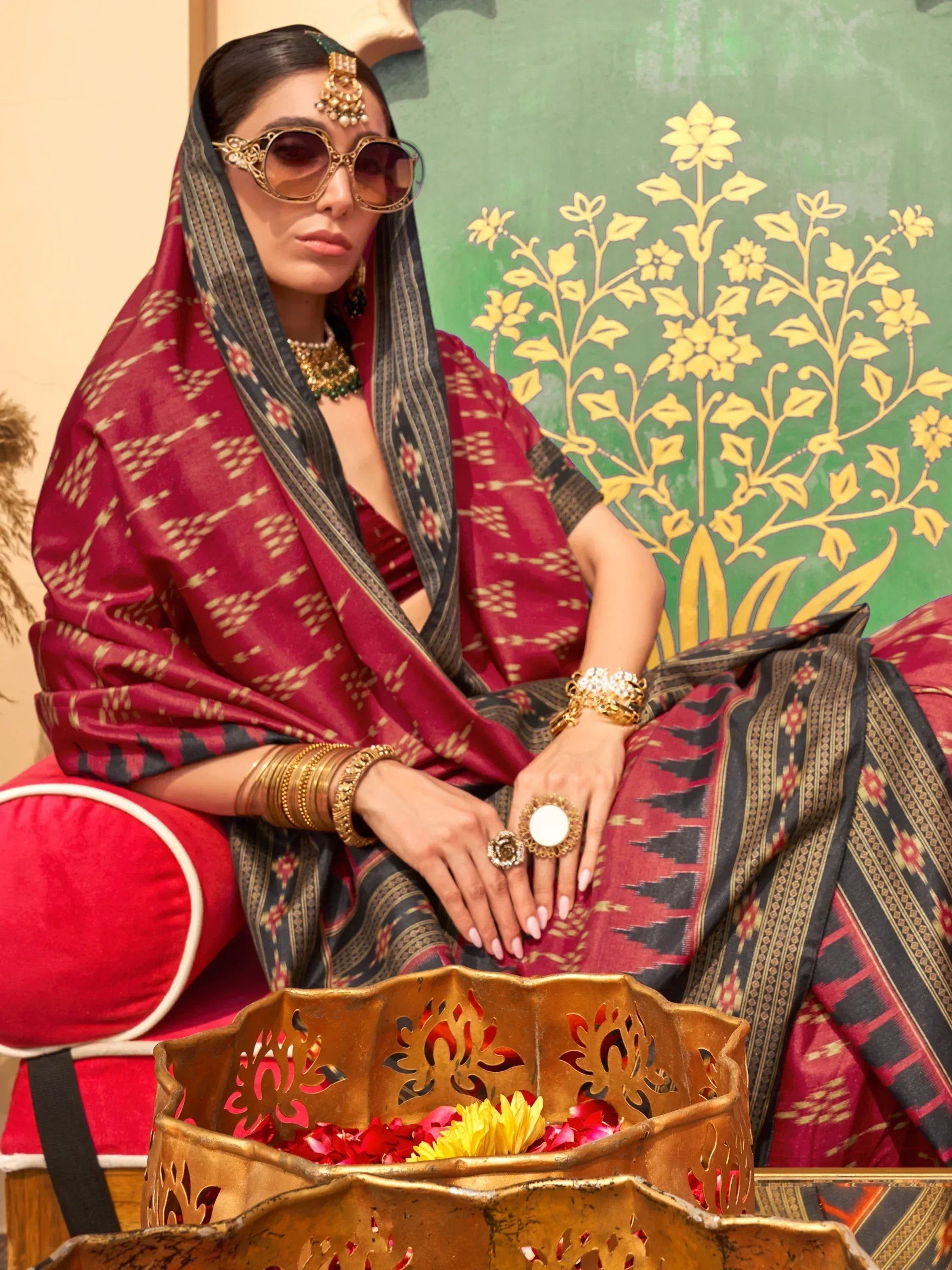 Woman in maroon and black ethnic saree with gold jewelry and sunglasses, seated with floral decor