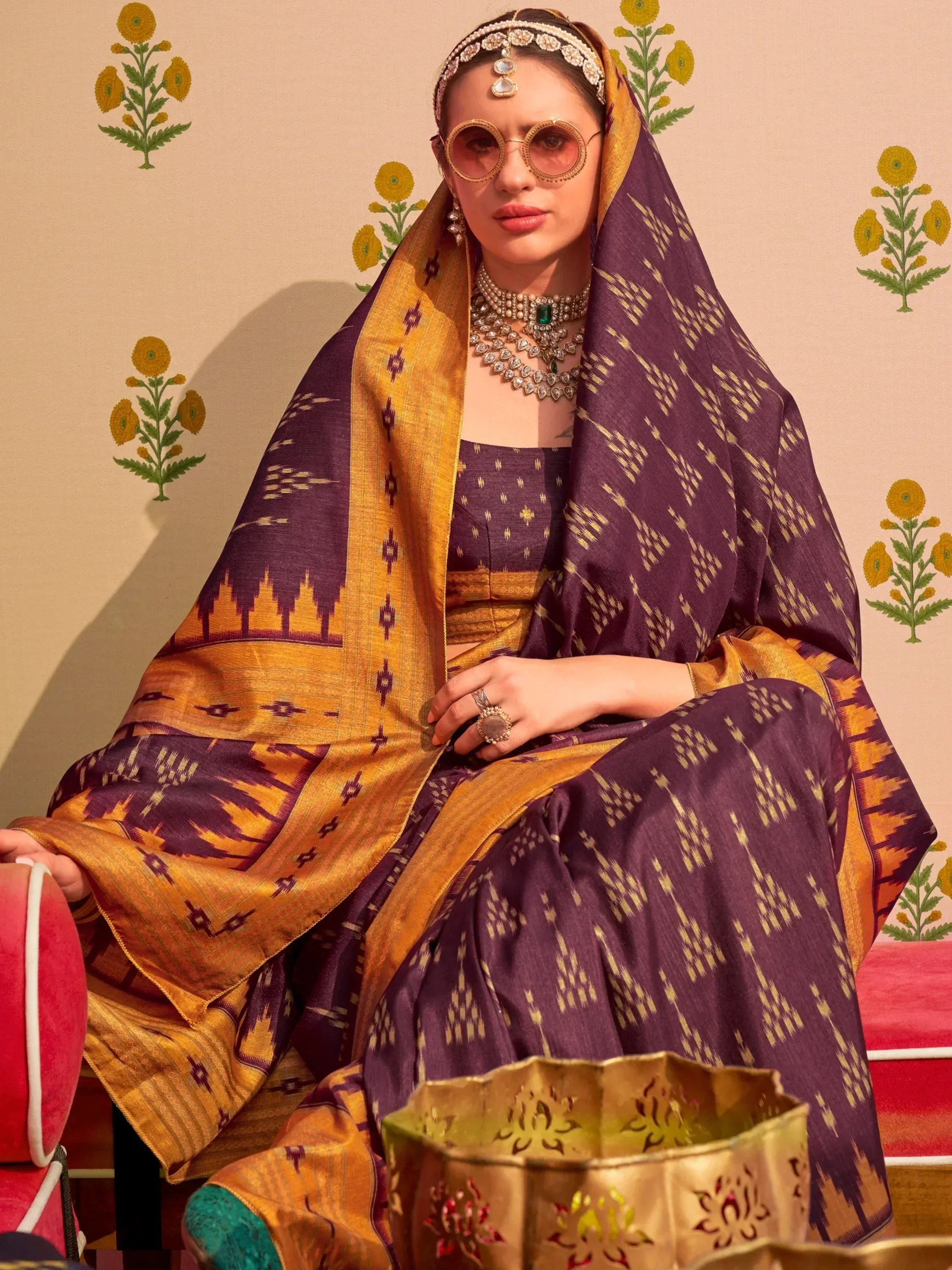 Woman in purple and gold ethnic saree with traditional jewelry and sunglasses, floral backdrop
