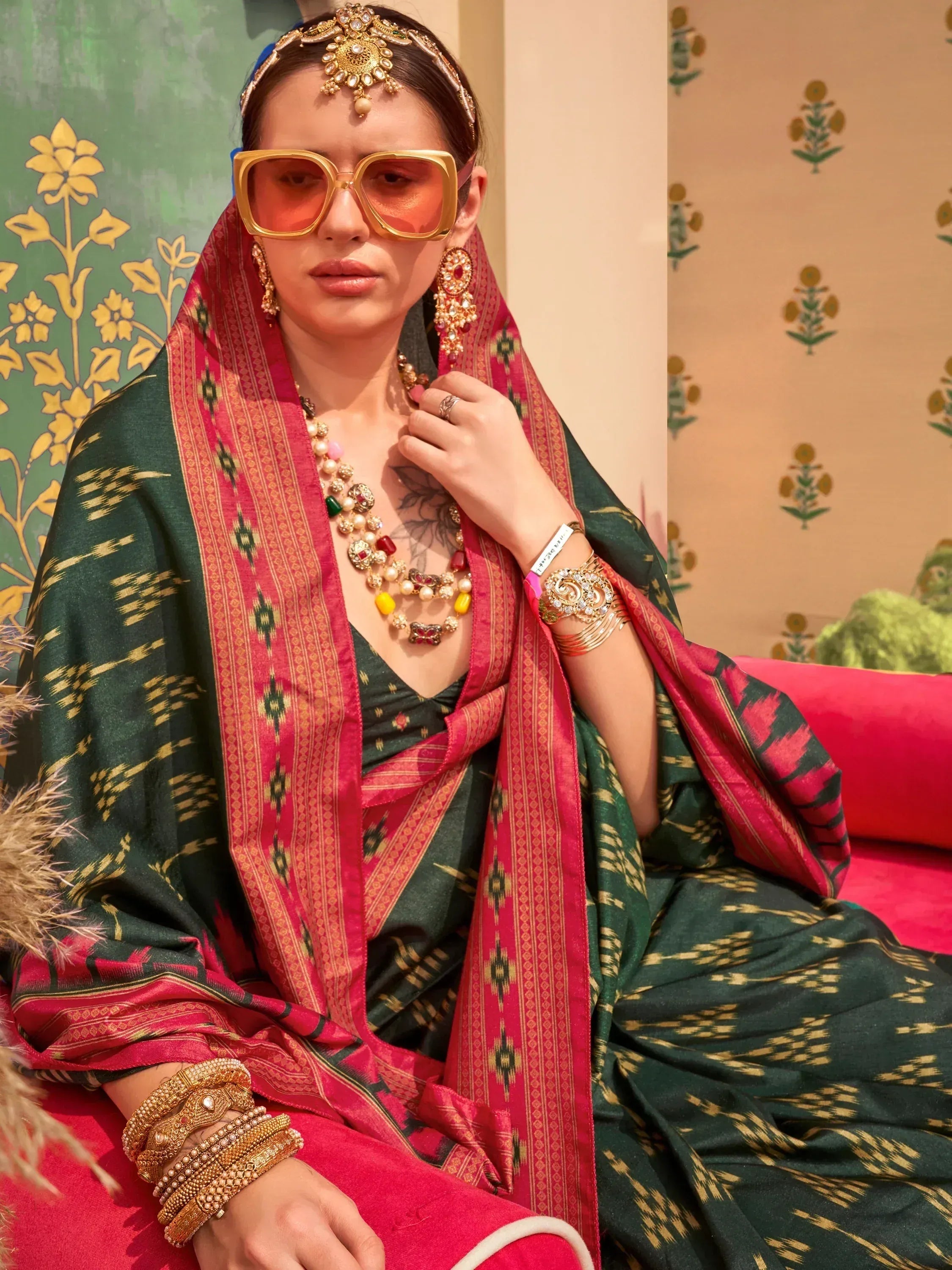 Woman in green and pink ethnic saree with gold jewelry and statement sunglasses, styled for festive occasions.