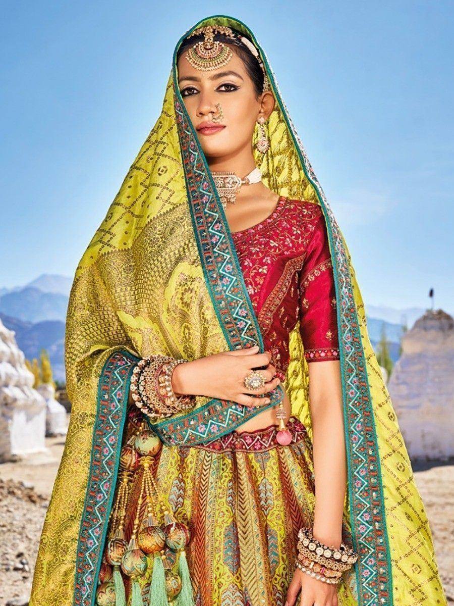Woman in vibrant red and yellow designer lehenga choli with intricate embroidery, traditional jewelry, and dupatta, set against mountain backdrop
