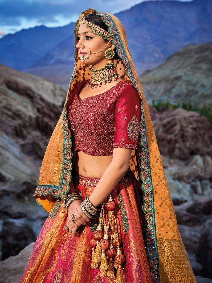 Indian bride in maroon lehenga choli with gold embroidery, orange dupatta, and traditional jewelry against mountain backdrop