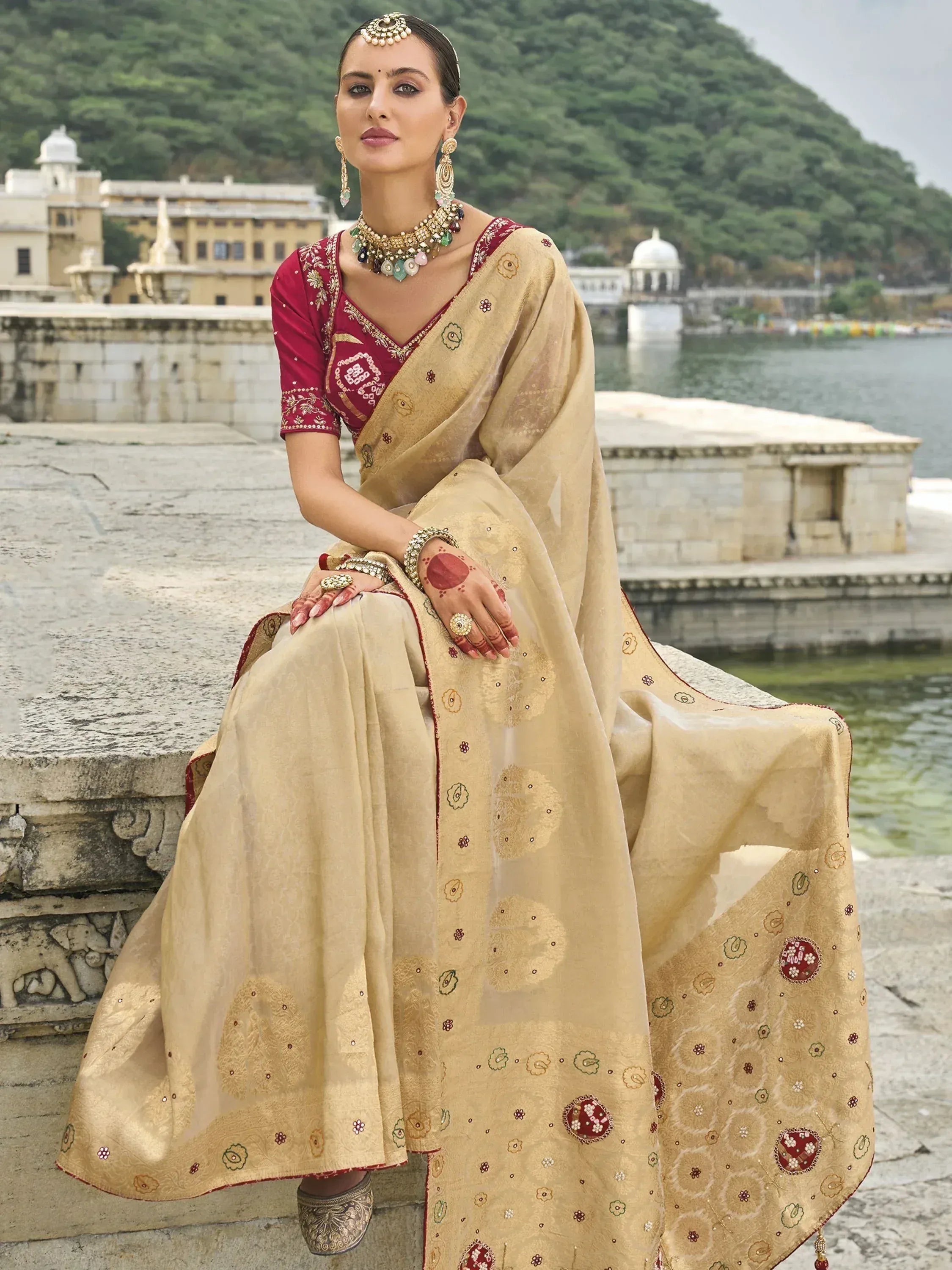 Elegant woman in beige and maroon embroidered saree with traditional jewelry, seated by a lakeside palace