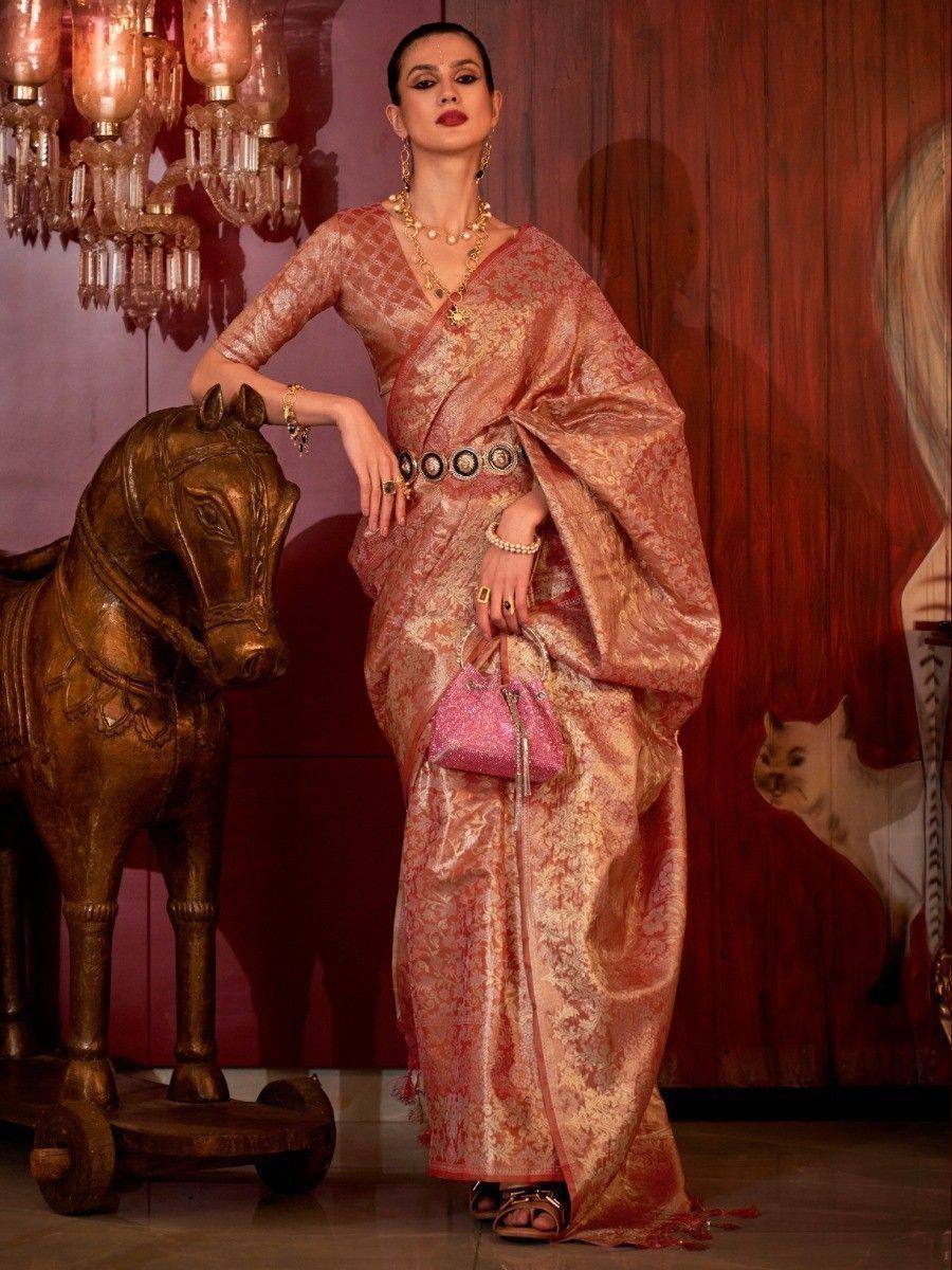 Elegant woman in designer pink and gold saree with intricate patterns, gold jewelry, and pink handbag, posing beside ornate horse statue.