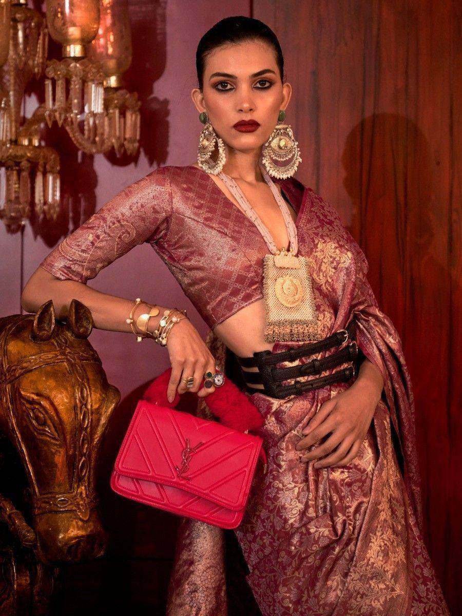 Woman in elegant maroon ethnic gown with gold embroidery, statement jewelry, and pink handbag, styled for festive occasions.