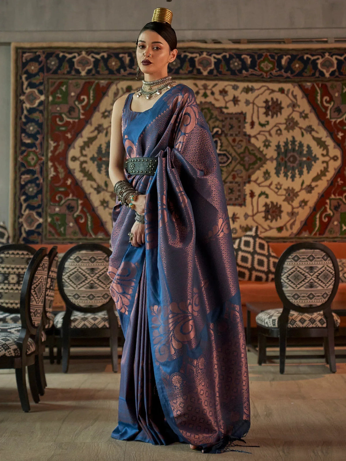 Woman in blue and purple printed saree with ethnic jewelry and gold headpiece, standing before ornate rug.