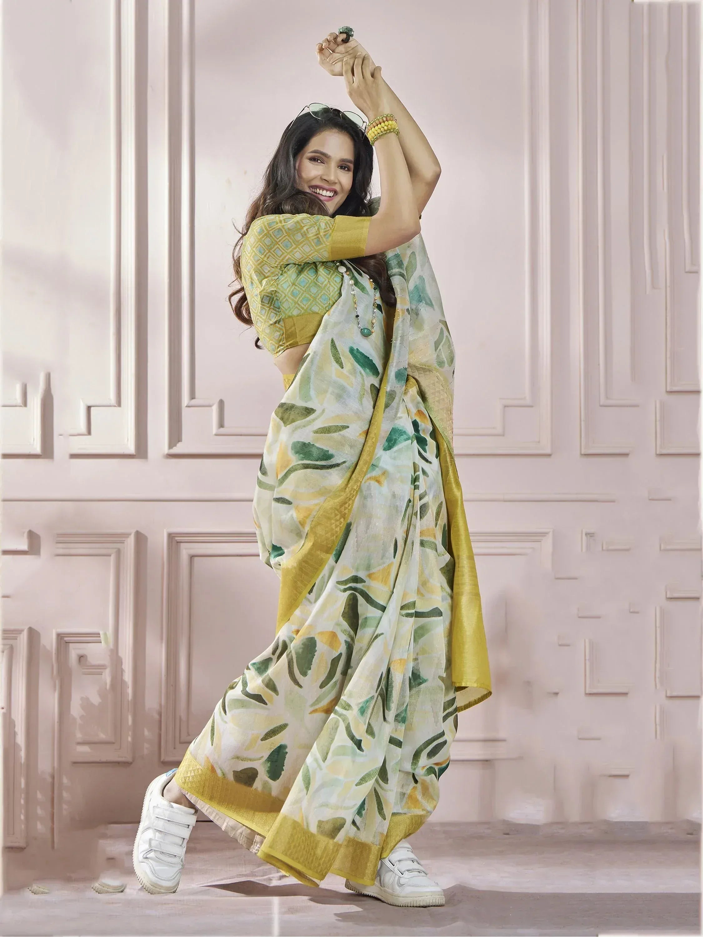 Woman in yellow green leaf print saree with matching blouse and white sneakers, posing against pink paneled wall