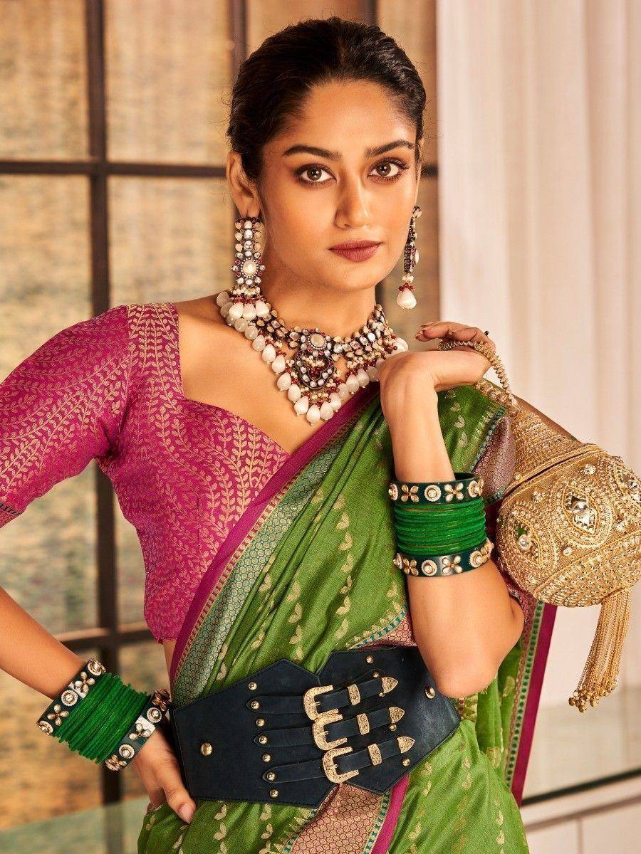 Woman in green saree with pink blouse, statement jewelry, and black belt, styled for ethnic occasions
