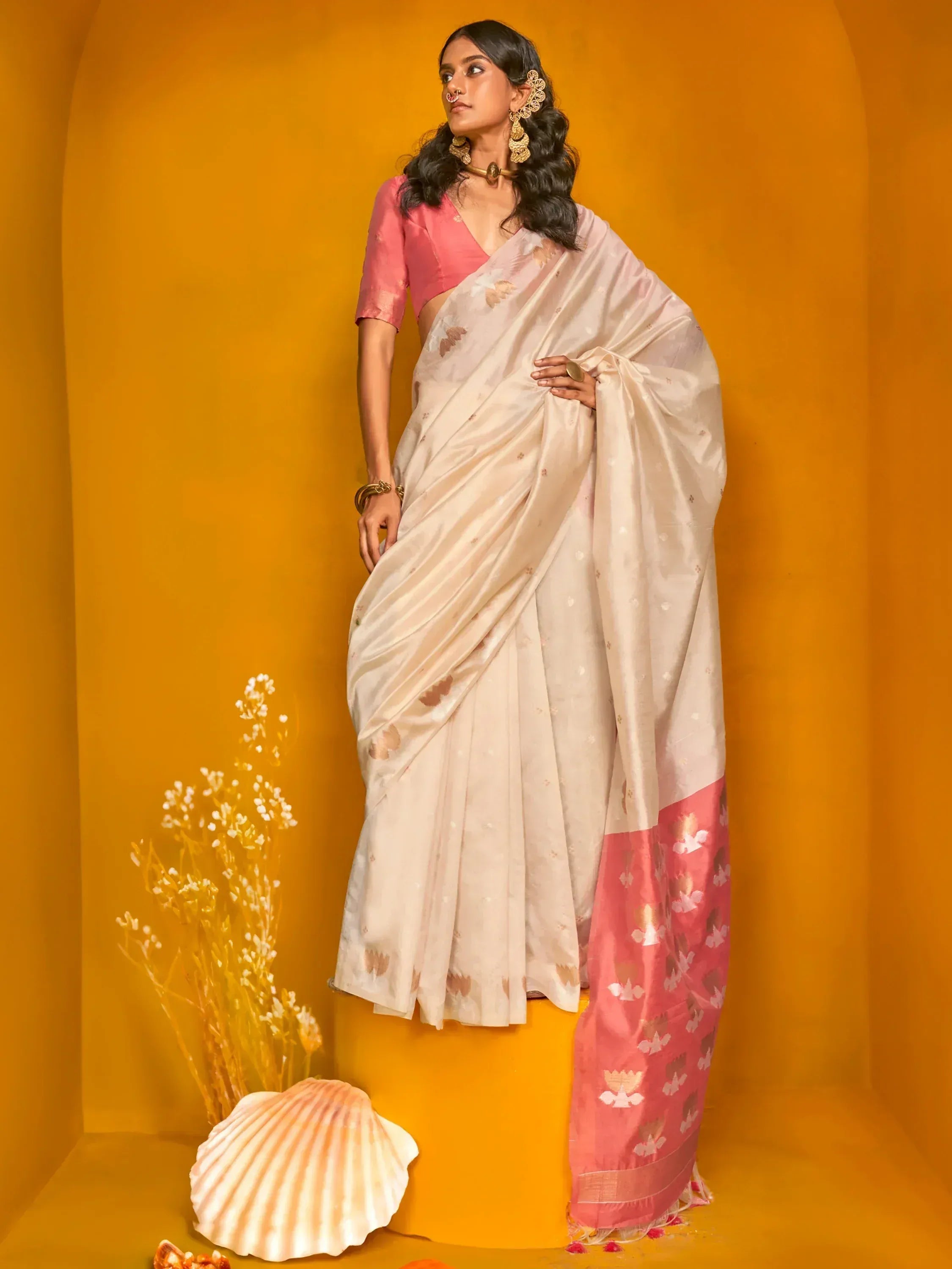 Woman in elegant cream saree with pink border and floral motifs, paired with pink blouse, gold jewelry, against warm orange backdrop.