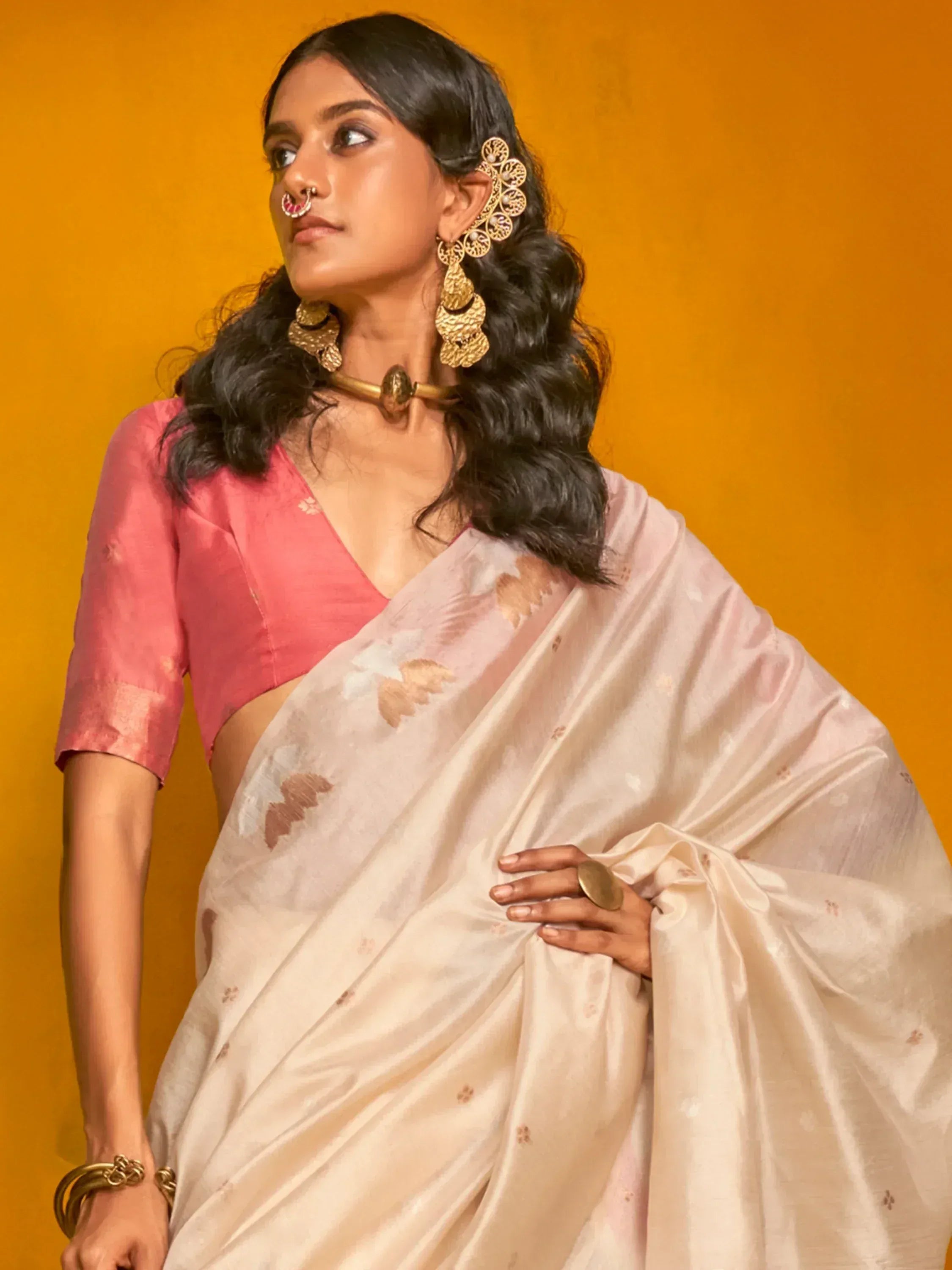 Woman in elegant cream saree with floral print, pink blouse, and gold jewelry against orange backdrop