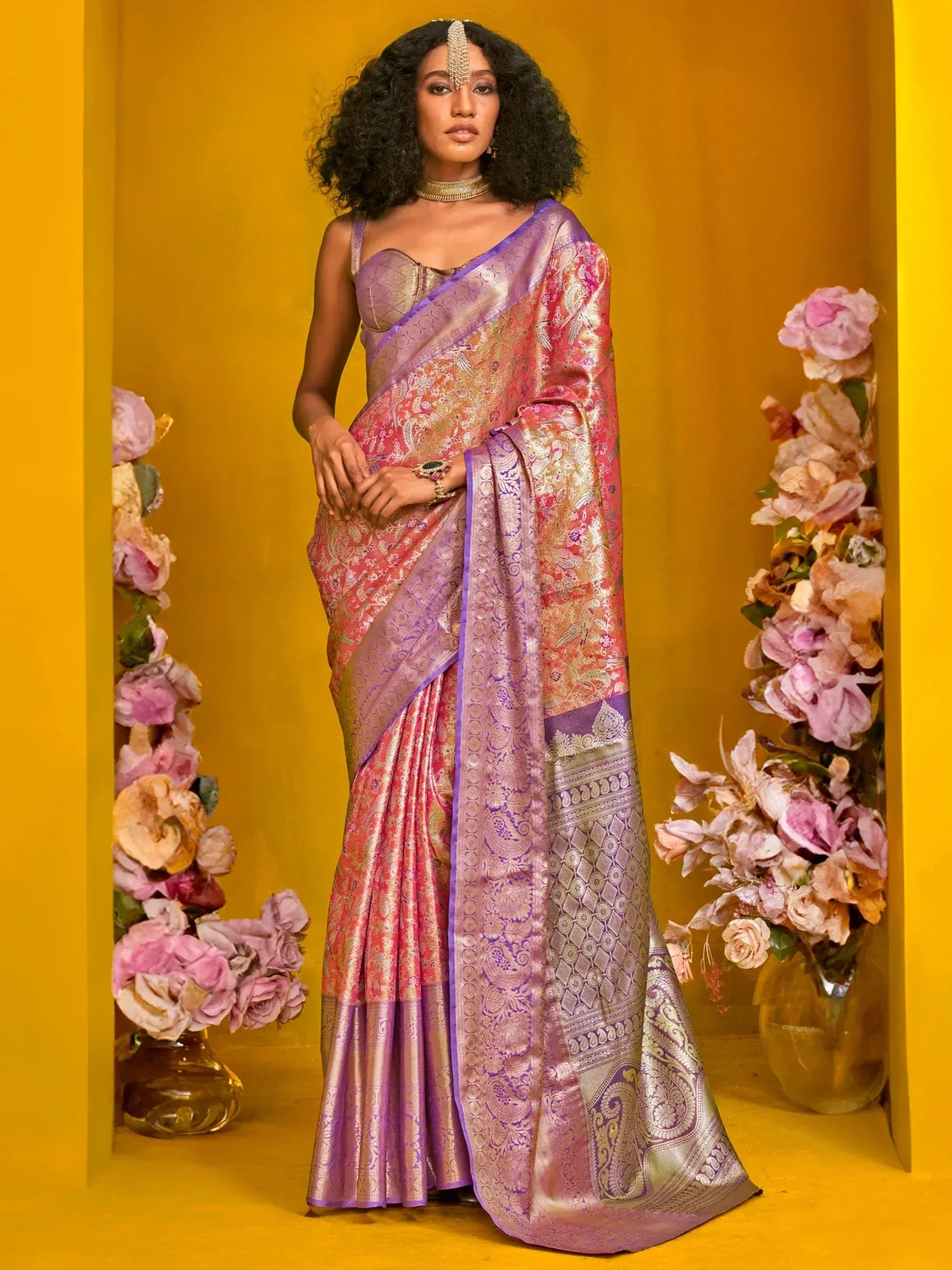 Elegant woman in designer purple and pink saree with floral patterns, styled with gold jewelry against a vibrant yellow backdrop.