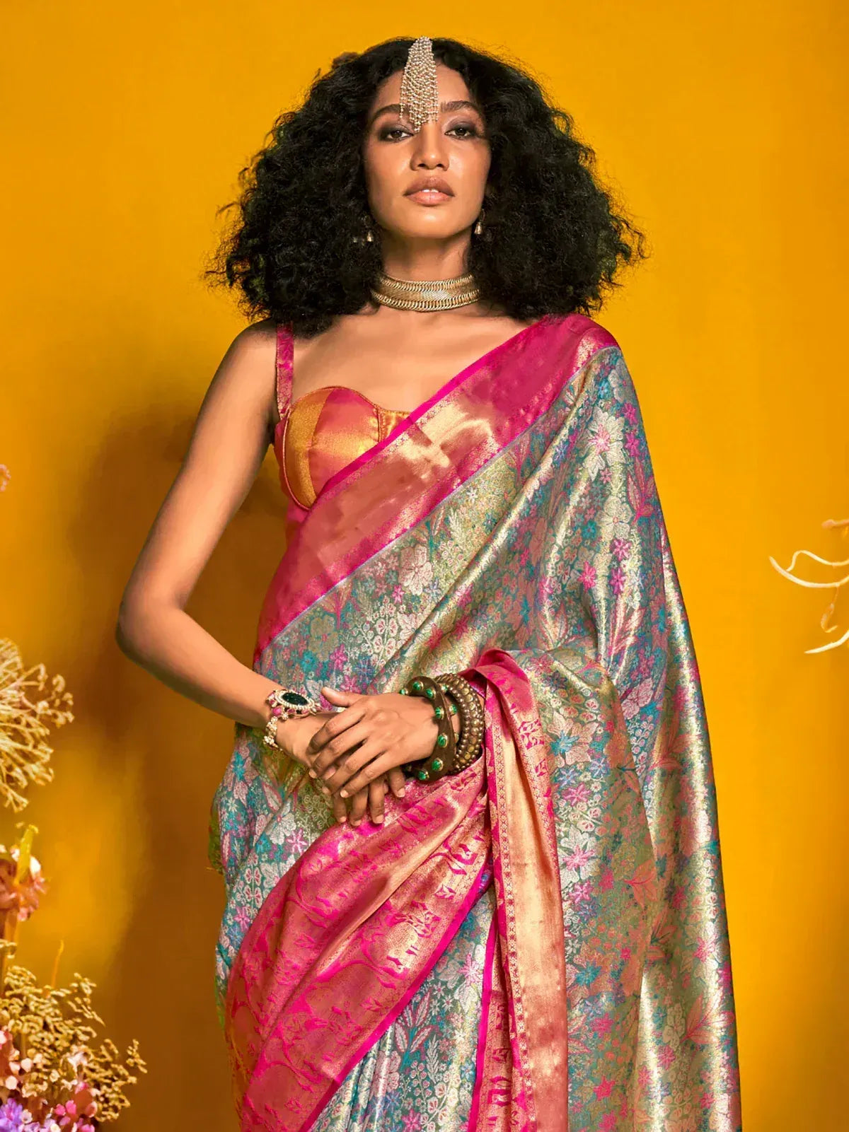 Elegant woman in vibrant pink and gold floral saree with traditional jewelry against yellow backdrop