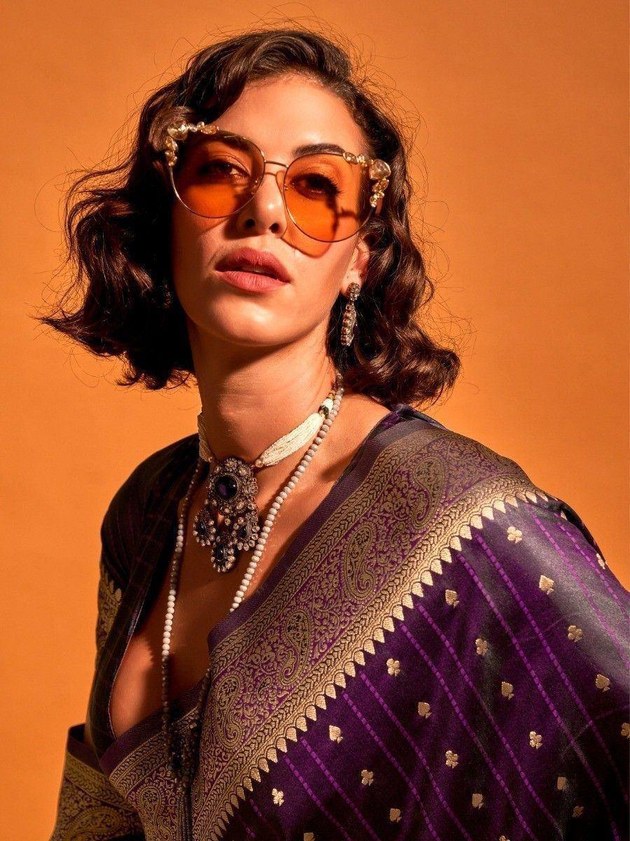 Woman in purple ethnic saree with gold paisley border, statement jewelry, and ornate sunglasses
