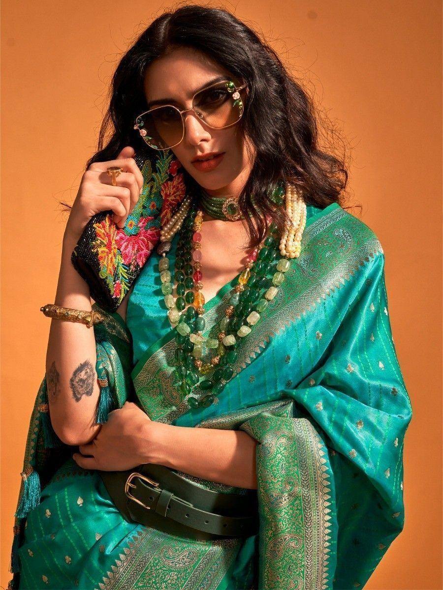 Elegant teal saree with intricate paisley designs, paired with layered gemstone necklaces and stylish sunglasses.
