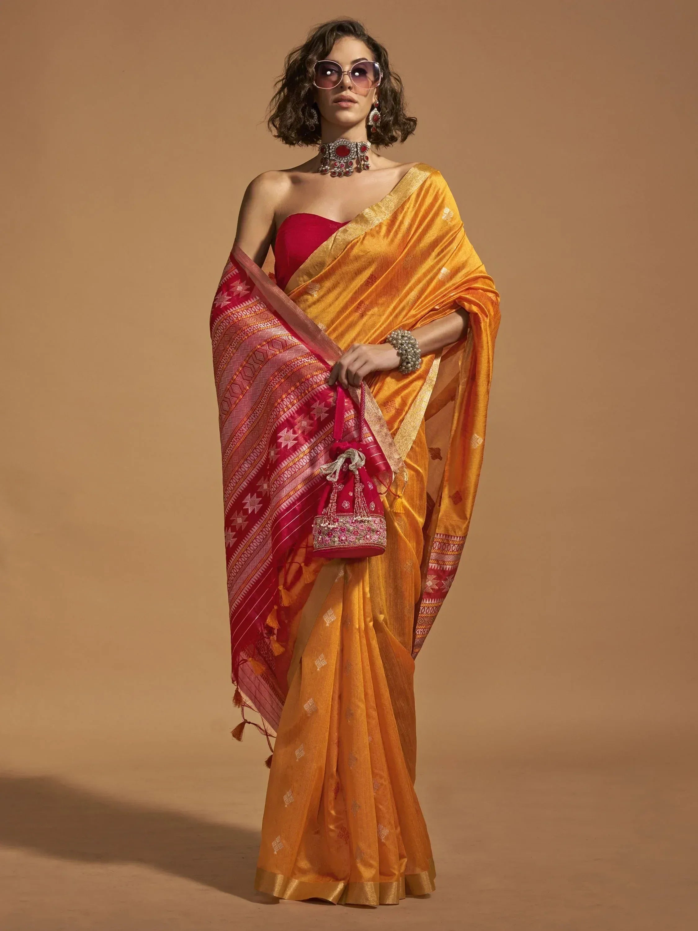 Woman in vibrant orange and red ethnic saree with gold border, paired with statement jewelry and matching potli bag.