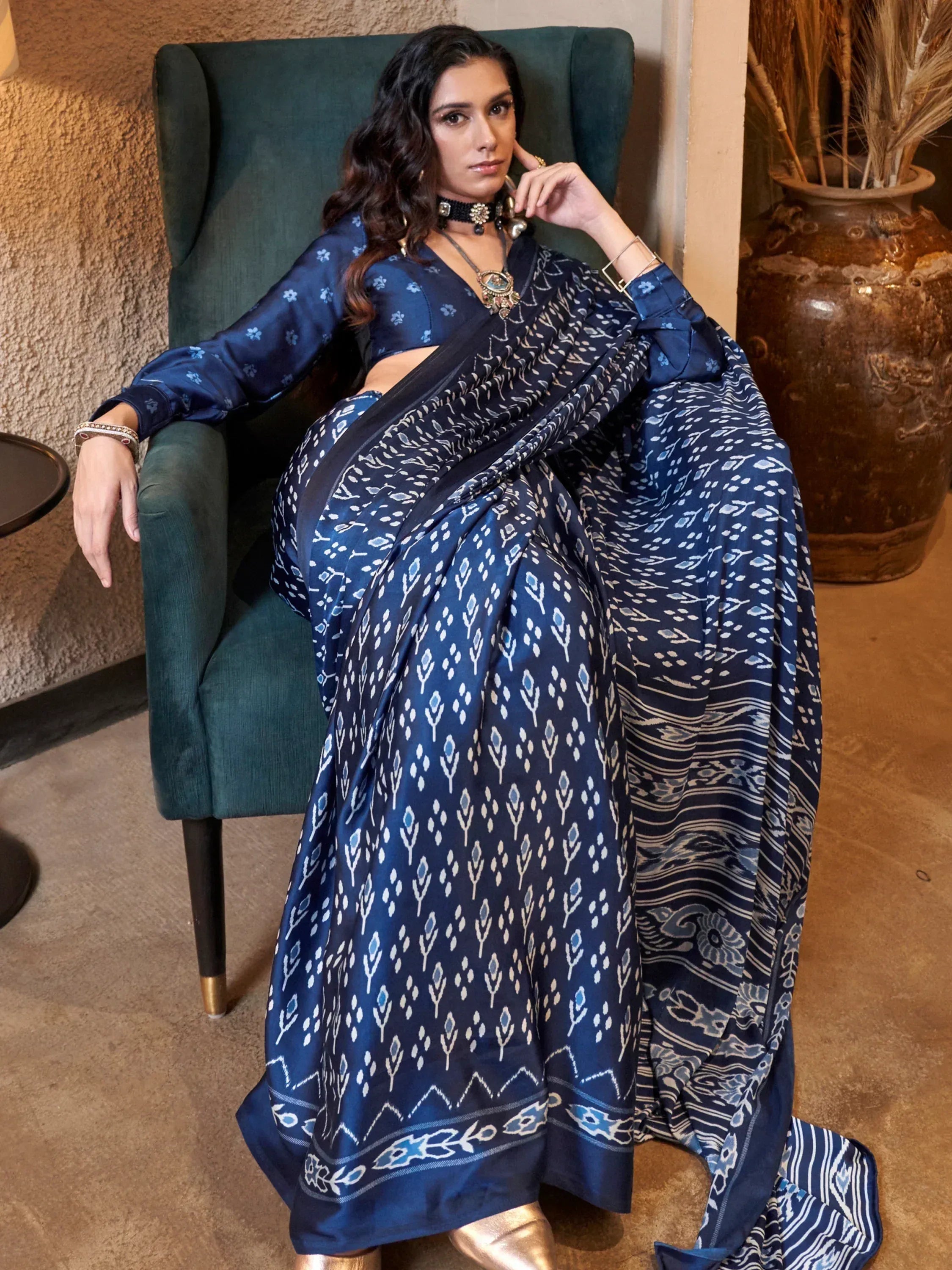 Woman in navy blue printed saree with matching blouse, ethnic jewelry, seated on green armchair