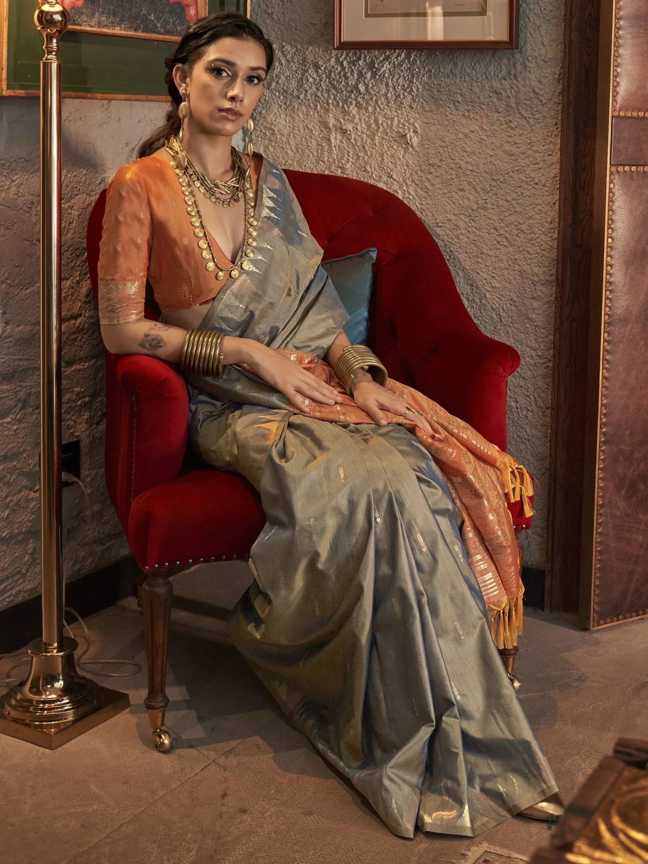 Woman in elegant grey and orange saree with gold jewelry, seated on red velvet chair