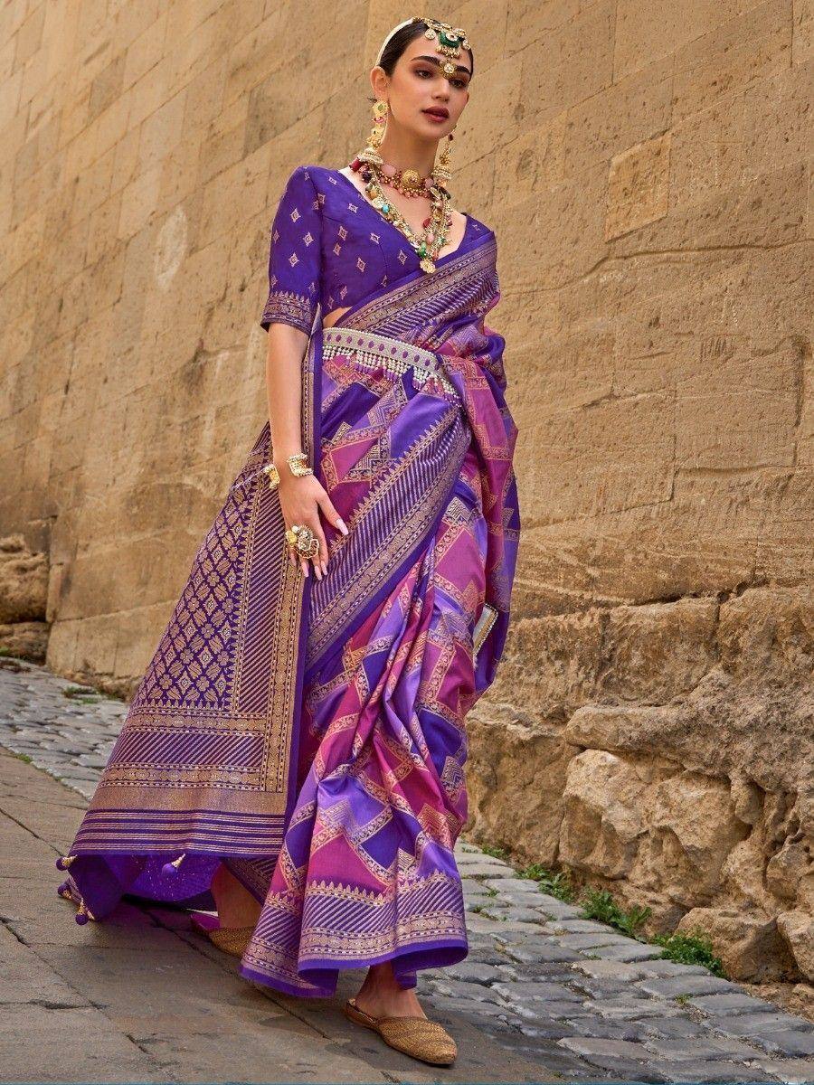 Purple and pink ethnic saree with gold embroidery, worn with traditional jewelry against stone wall