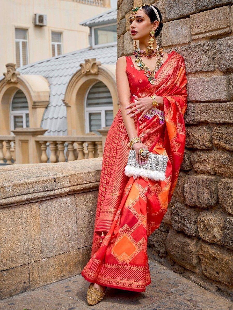 Woman in vibrant red and orange ethnic saree with gold jewelry, standing by stone wall