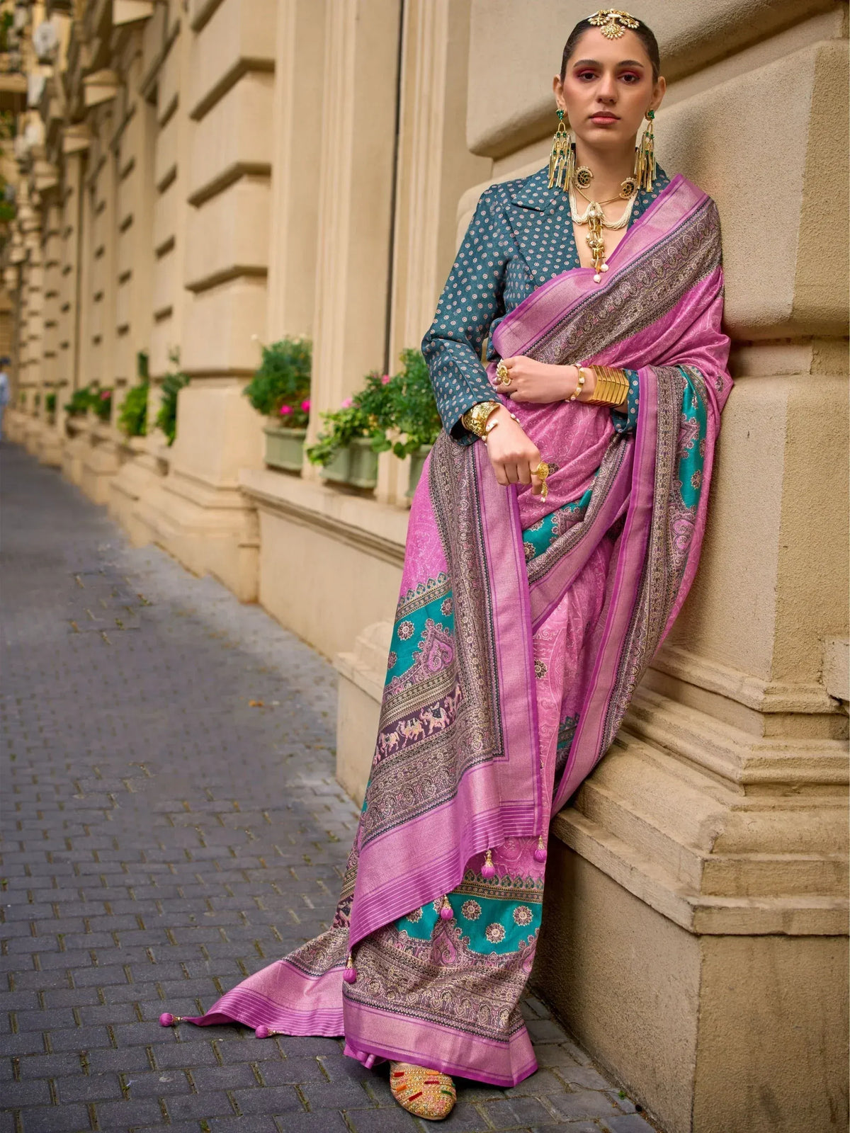 Woman in pink and teal designer saree with gold jewelry, leaning against stone wall