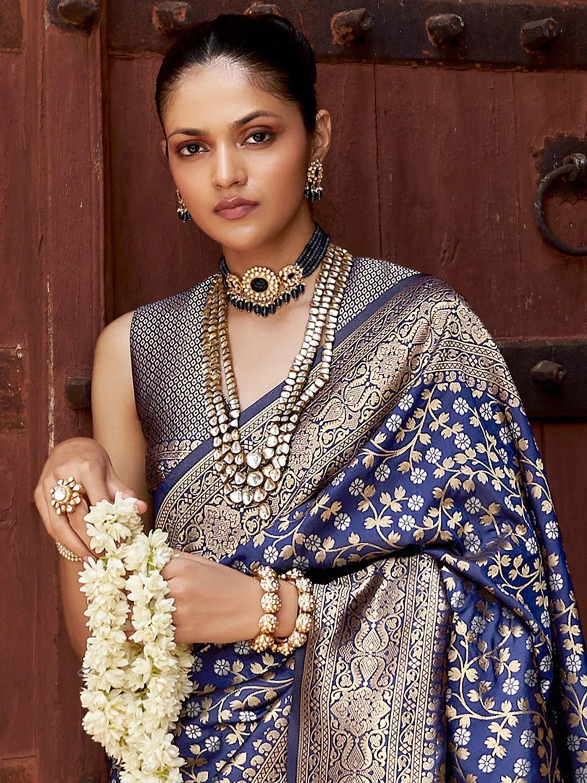 Elegant woman in blue and gold embroidered saree with traditional jewelry, holding white flowers against rustic door