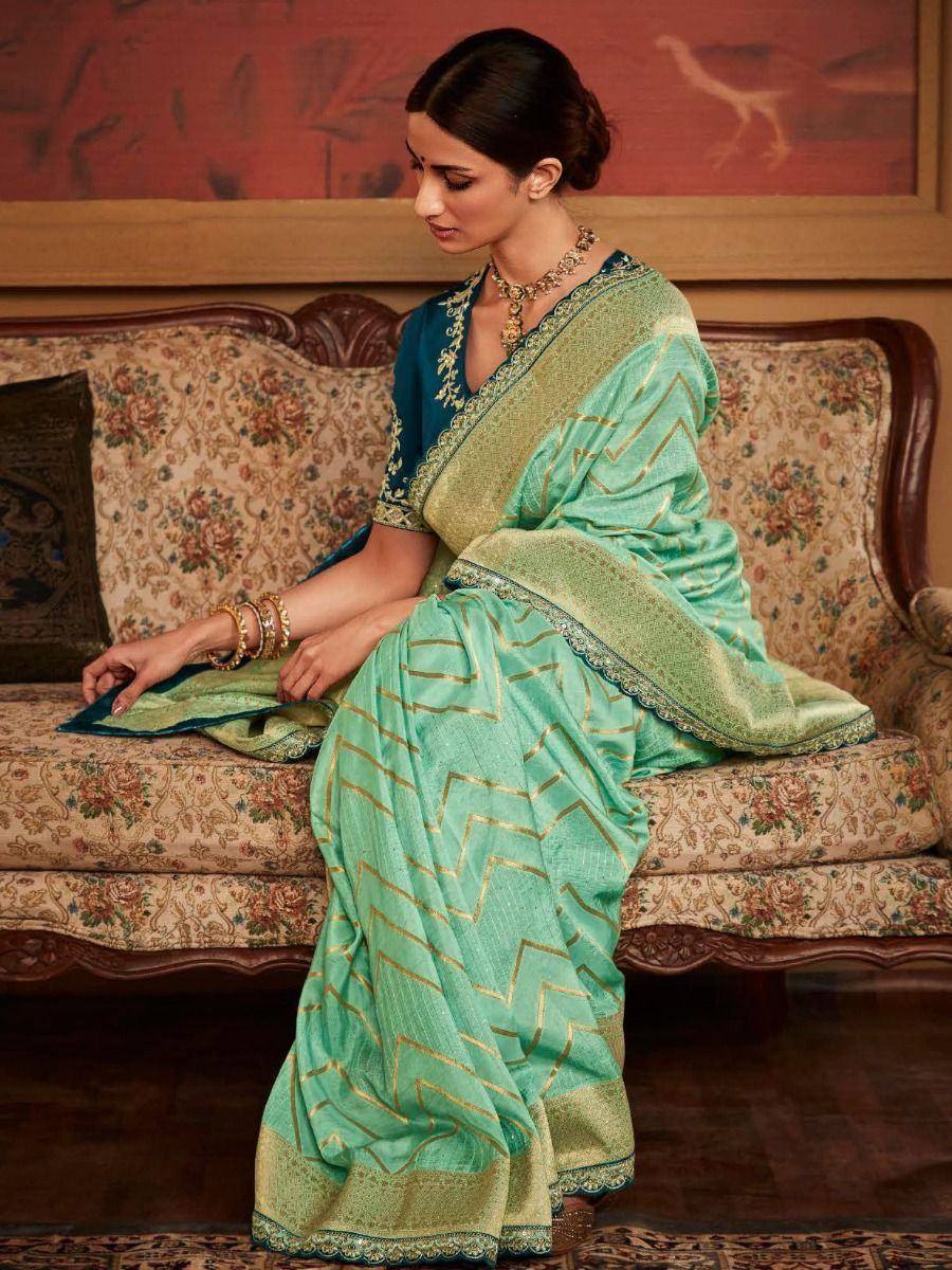 Woman in mint green saree with gold zigzag print, paired with blue blouse and traditional jewelry, seated on floral sofa