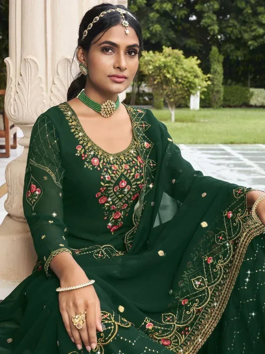 Woman in dark green embroidered ethnic gown with floral details, wearing traditional jewelry, seated outdoors.