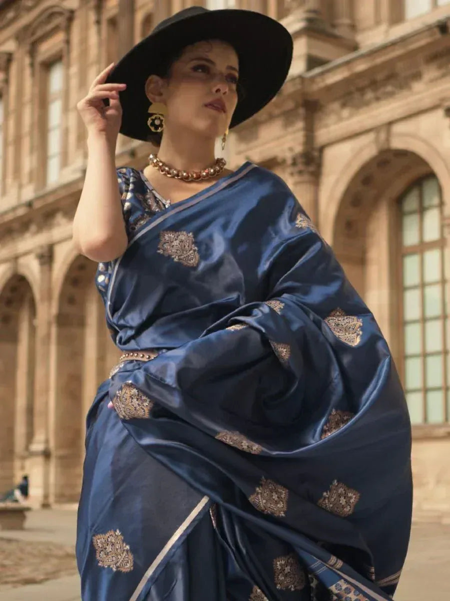 Woman in elegant blue silk saree with gold embroidery, styled with black hat and statement jewelry