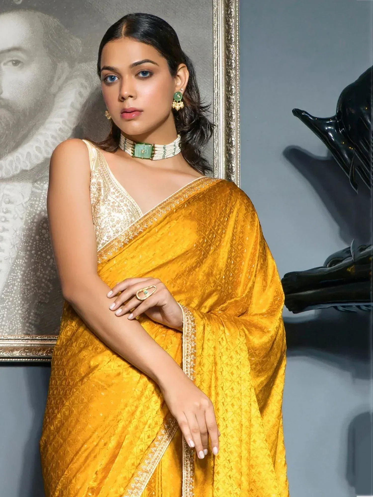 Woman in elegant yellow designer saree with gold embroidery, paired with green jewelry, perfect for ethnic occasions.