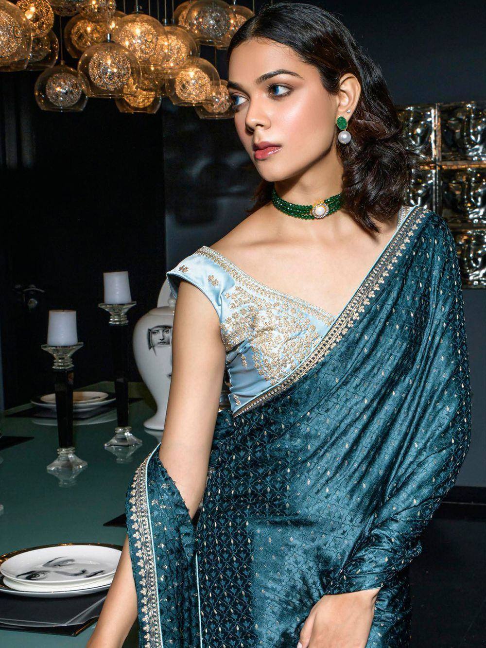 Elegant teal saree with gold embroidery and light blue blouse, styled with green jewelry for ethnic wear.