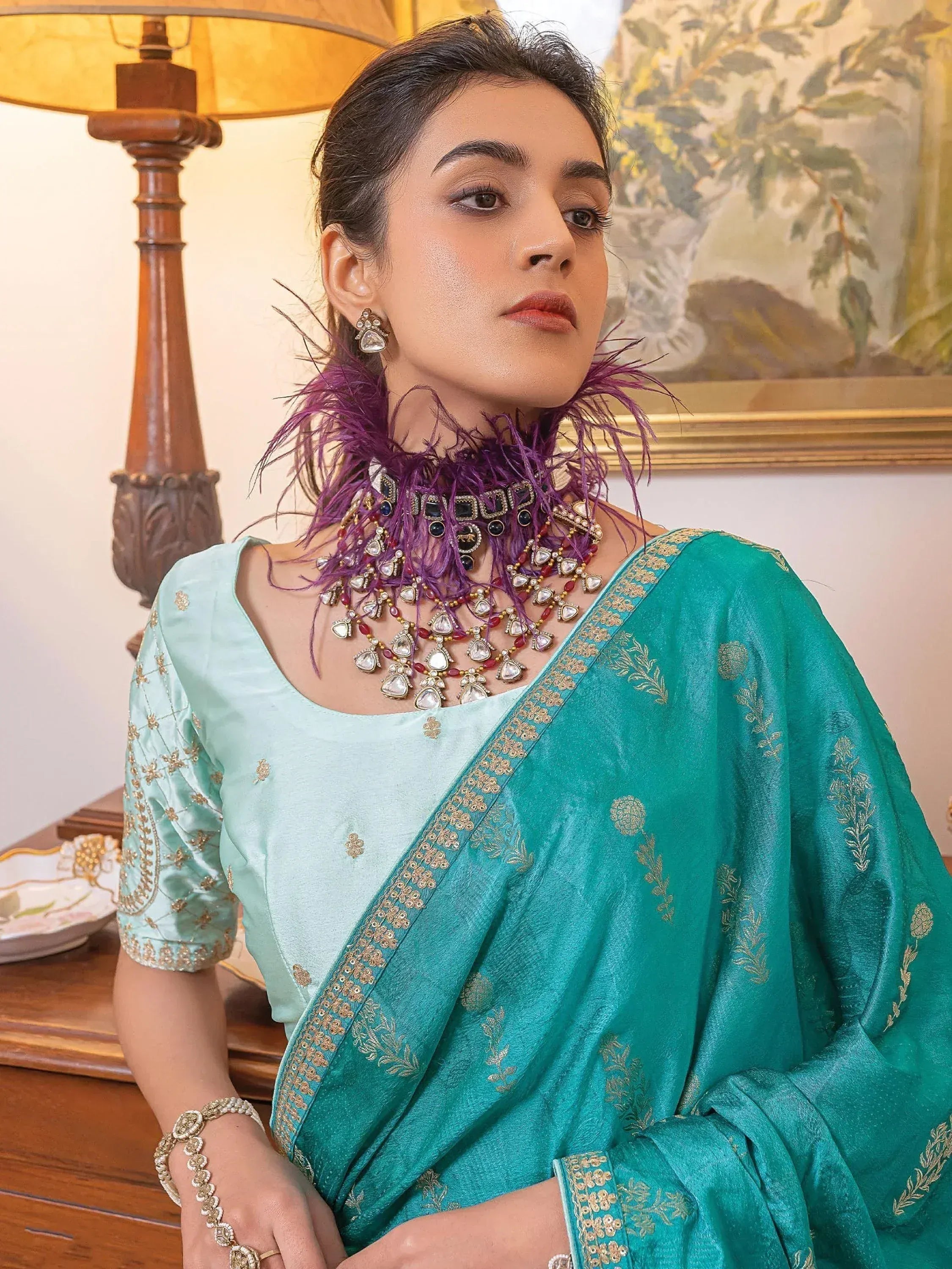 Woman in teal embroidered saree with gold detailing, paired with statement purple feather jewelry and matching blouse.
