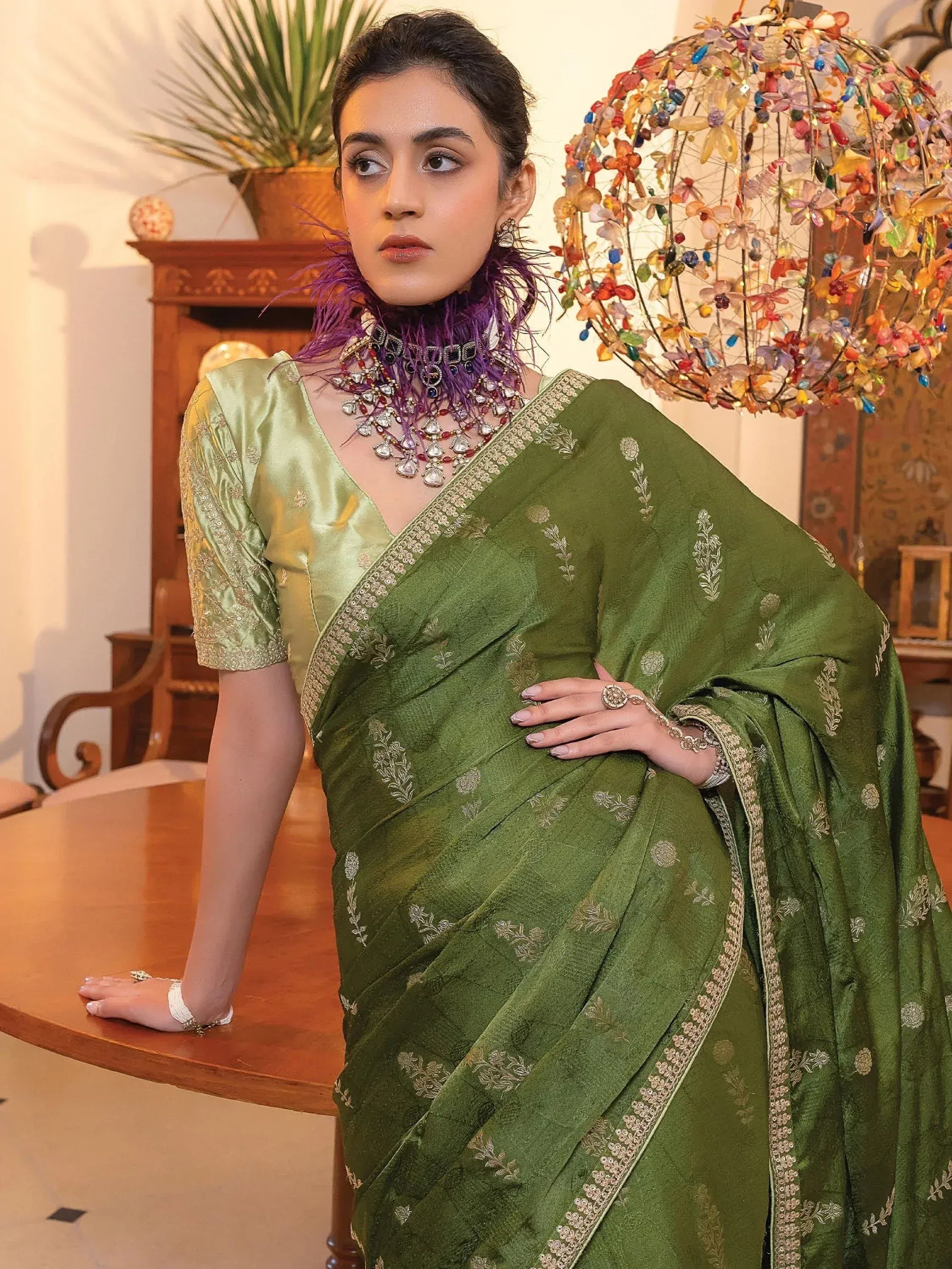 Elegant green embroidered saree with gold detailing, paired with statement jewelry and purple feather accents.