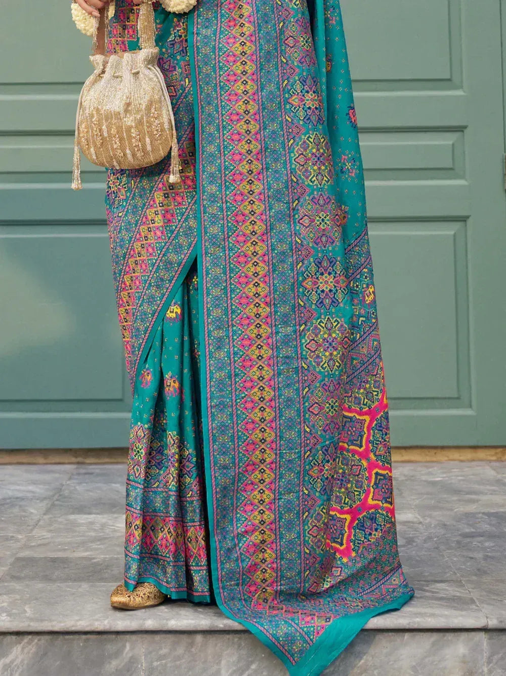 Teal printed saree with pink and gold geometric patterns, paired with a sequined potli bag and ethnic flats