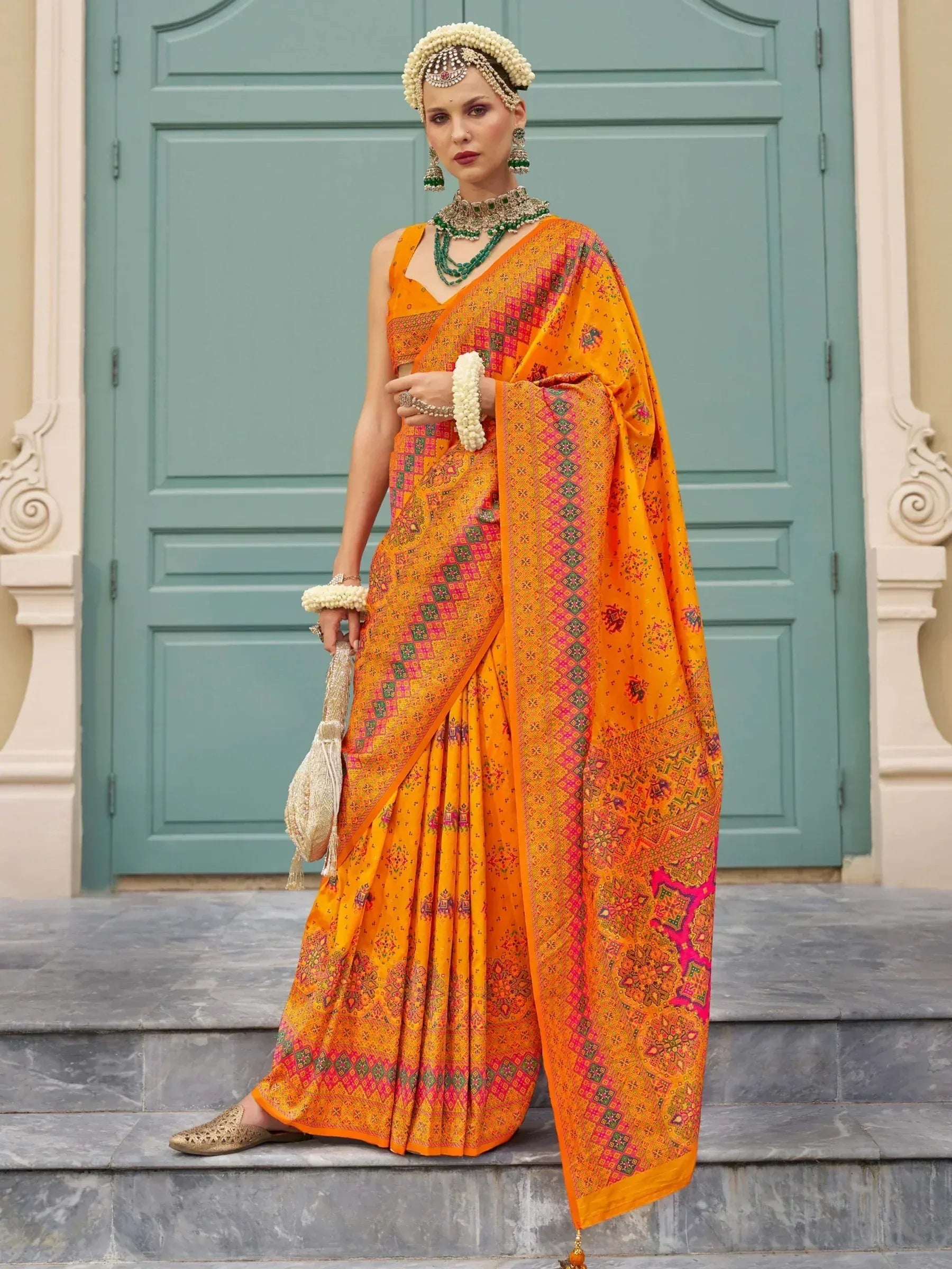 Vibrant orange ethnic saree with pink and green patterns, paired with traditional jewelry and floral headpiece.