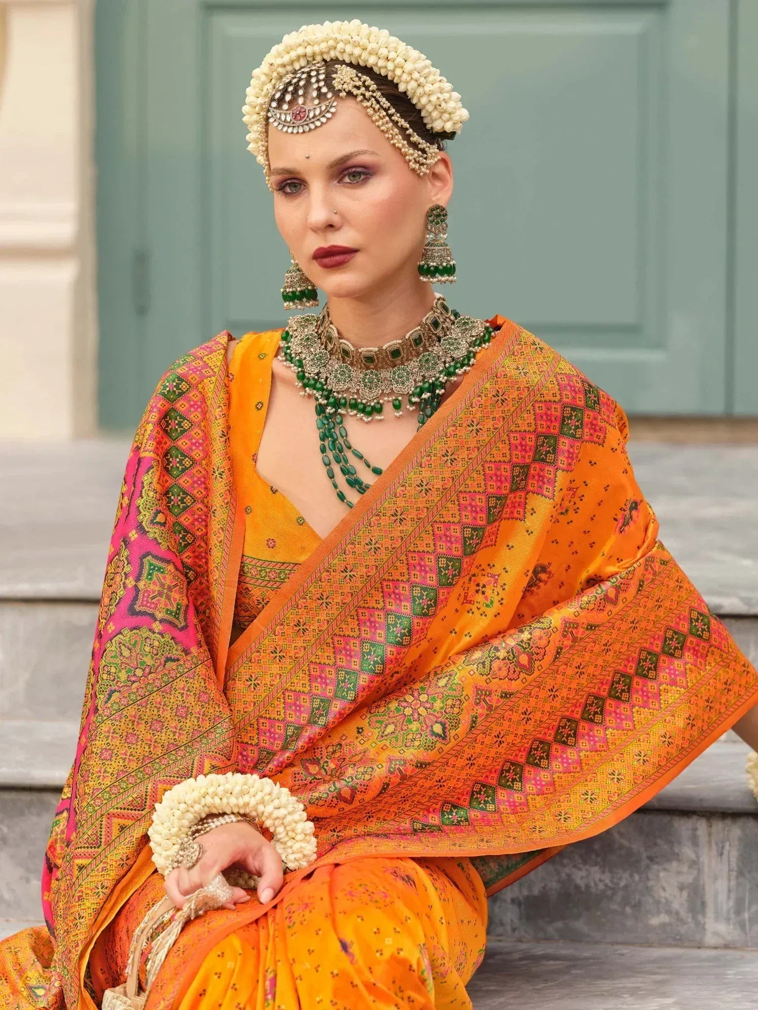Vibrant orange ethnic saree with pink and green geometric print, paired with traditional jewelry and floral headpiece.