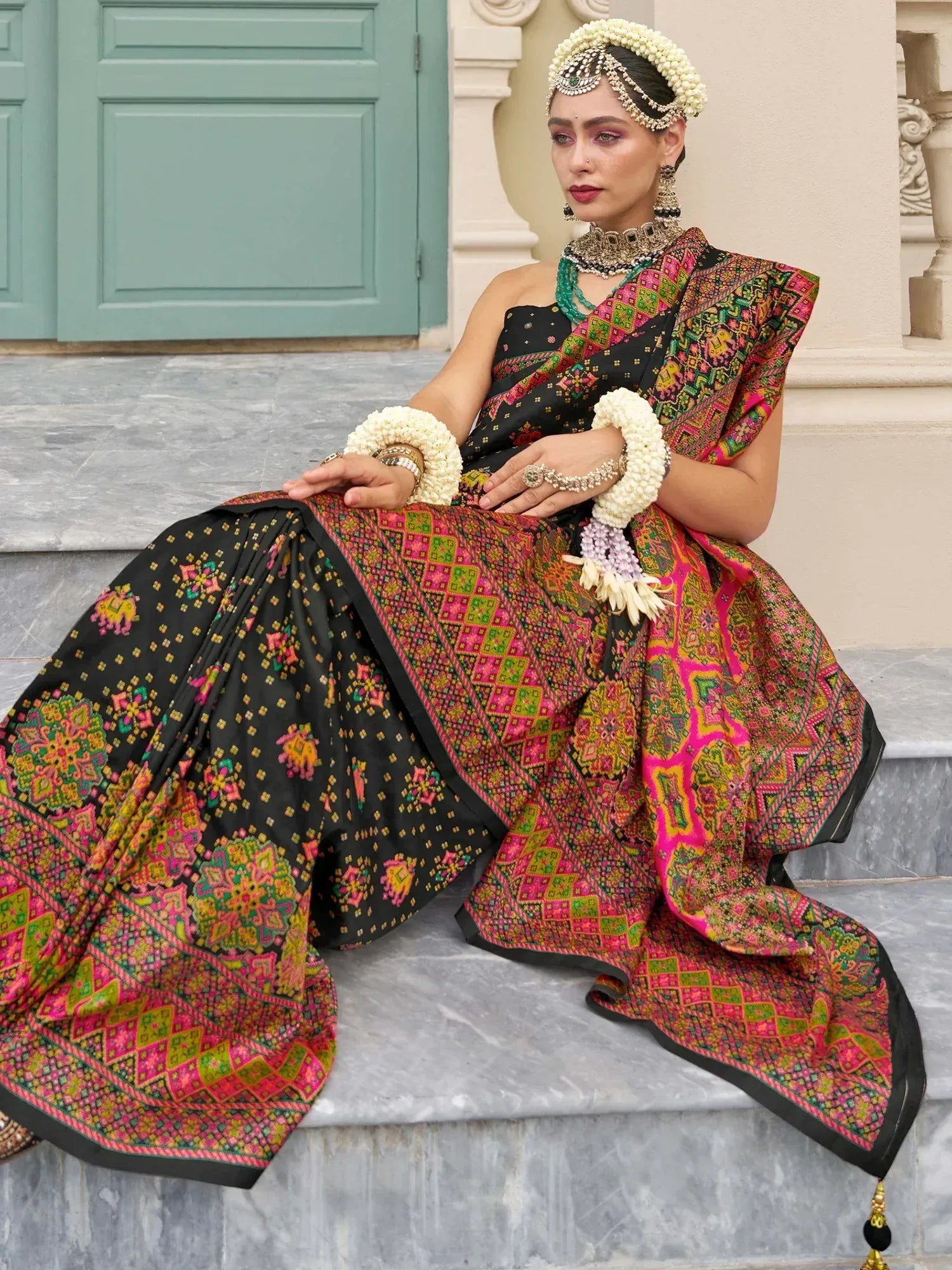Woman in vibrant black and pink embroidered lehenga choli with floral jewelry, seated on steps