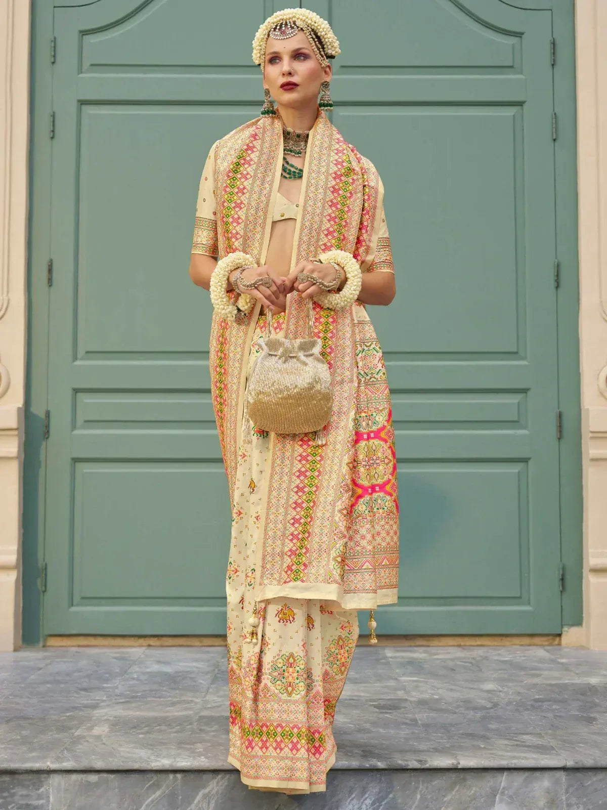 Woman in embroidered Indian ethnic wear with floral dupatta, pearl jewelry, and potli bag, standing before green door