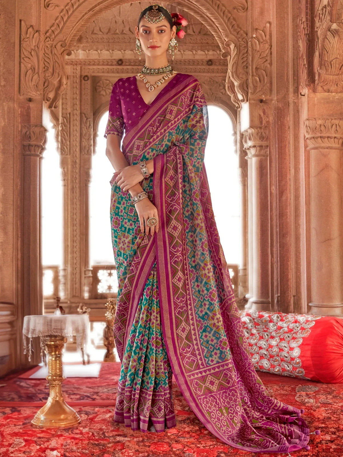 Woman in vibrant purple and teal ethnic saree with intricate geometric patterns, traditional jewelry, and floral hair accessory.