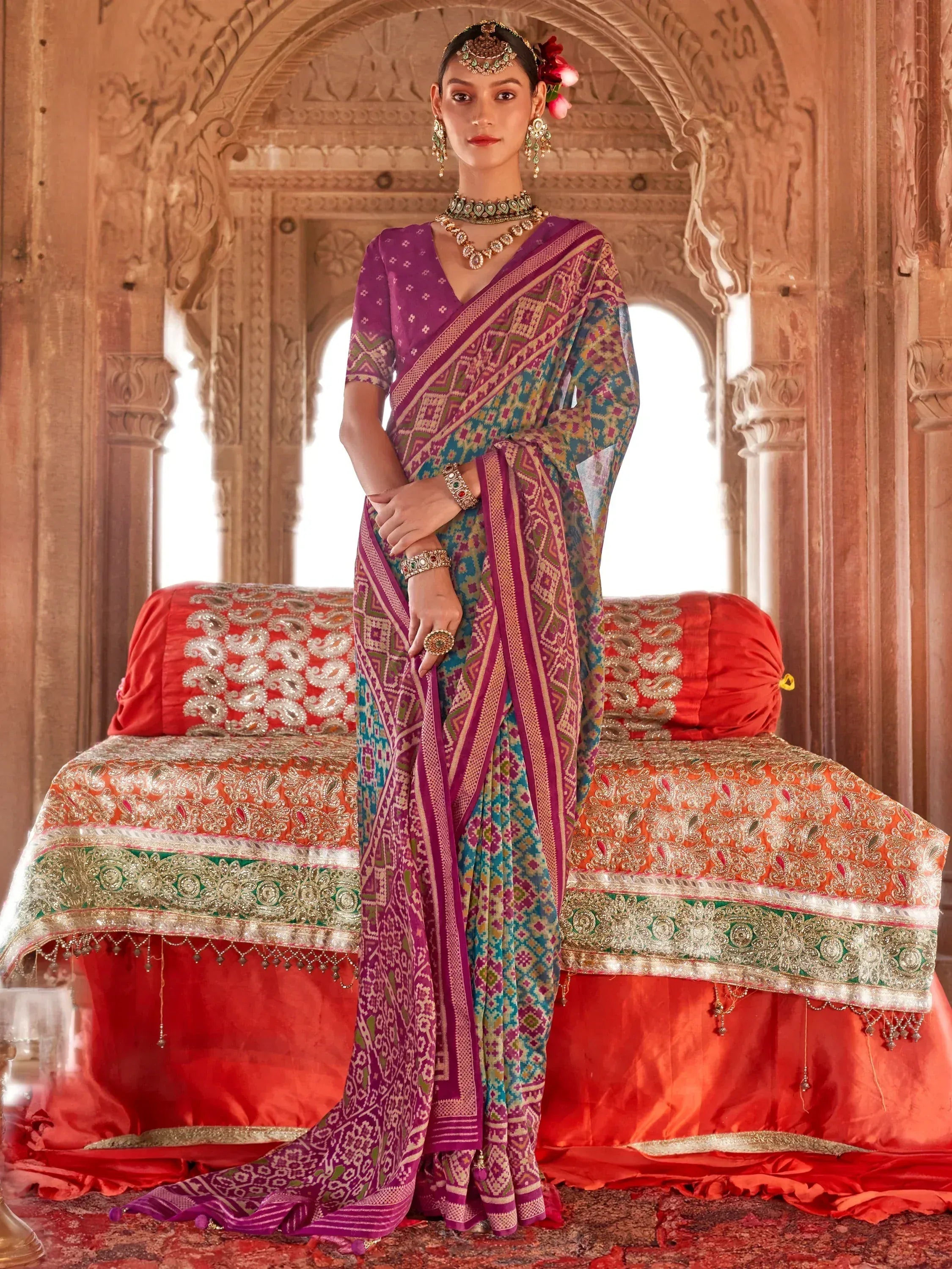 Woman in vibrant purple and teal printed saree with traditional jewelry, standing in ornate Indian palace setting