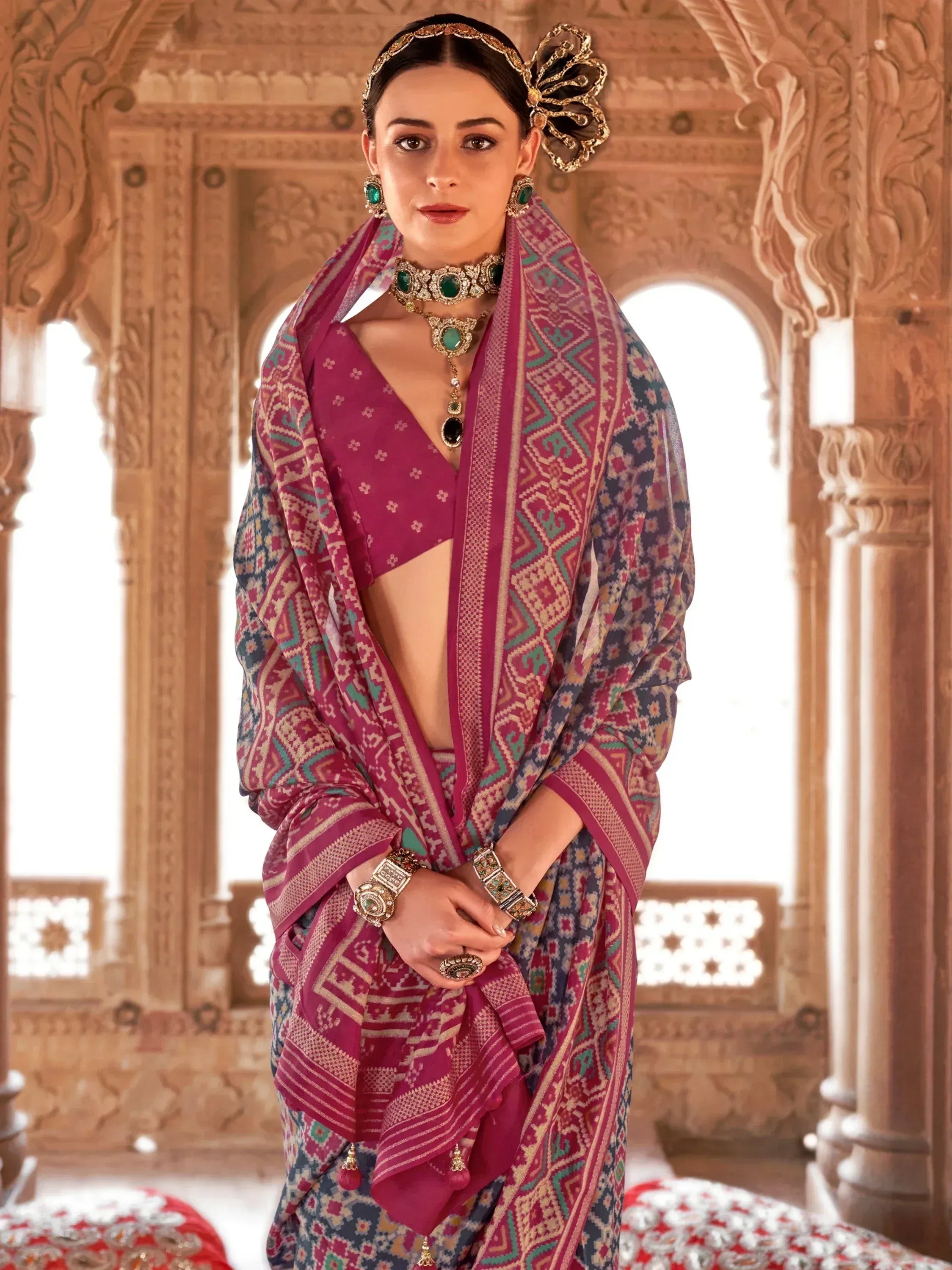 Woman in vibrant pink ethnic saree with geometric print, traditional jewelry, and ornate headpiece, set against carved palace backdrop.