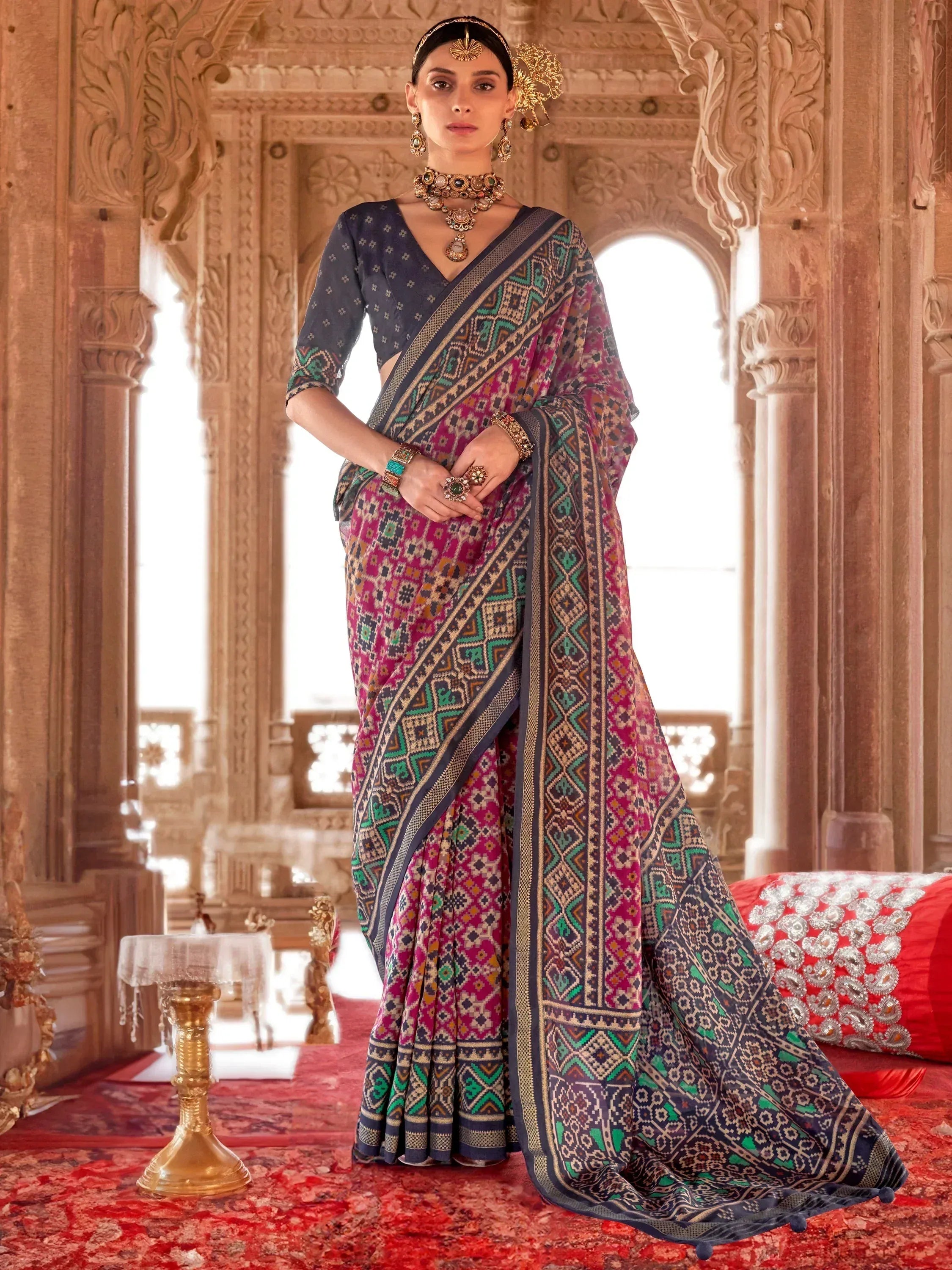 Woman in vibrant printed saree with ethnic jewelry, standing in ornate palace setting
