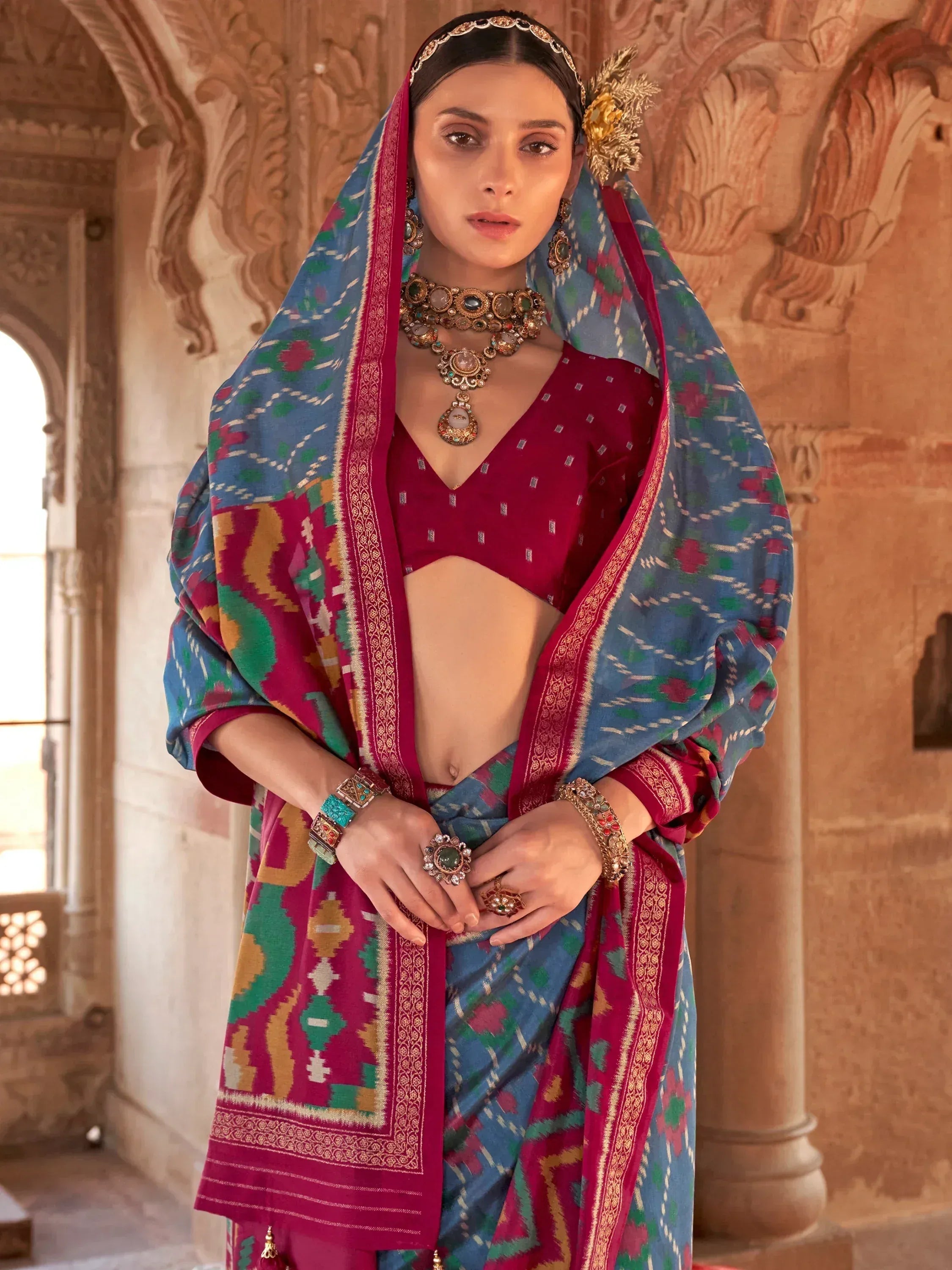 Woman in vibrant Indian ethnic wear with patterned dupatta, traditional jewelry, and ornate palace backdrop